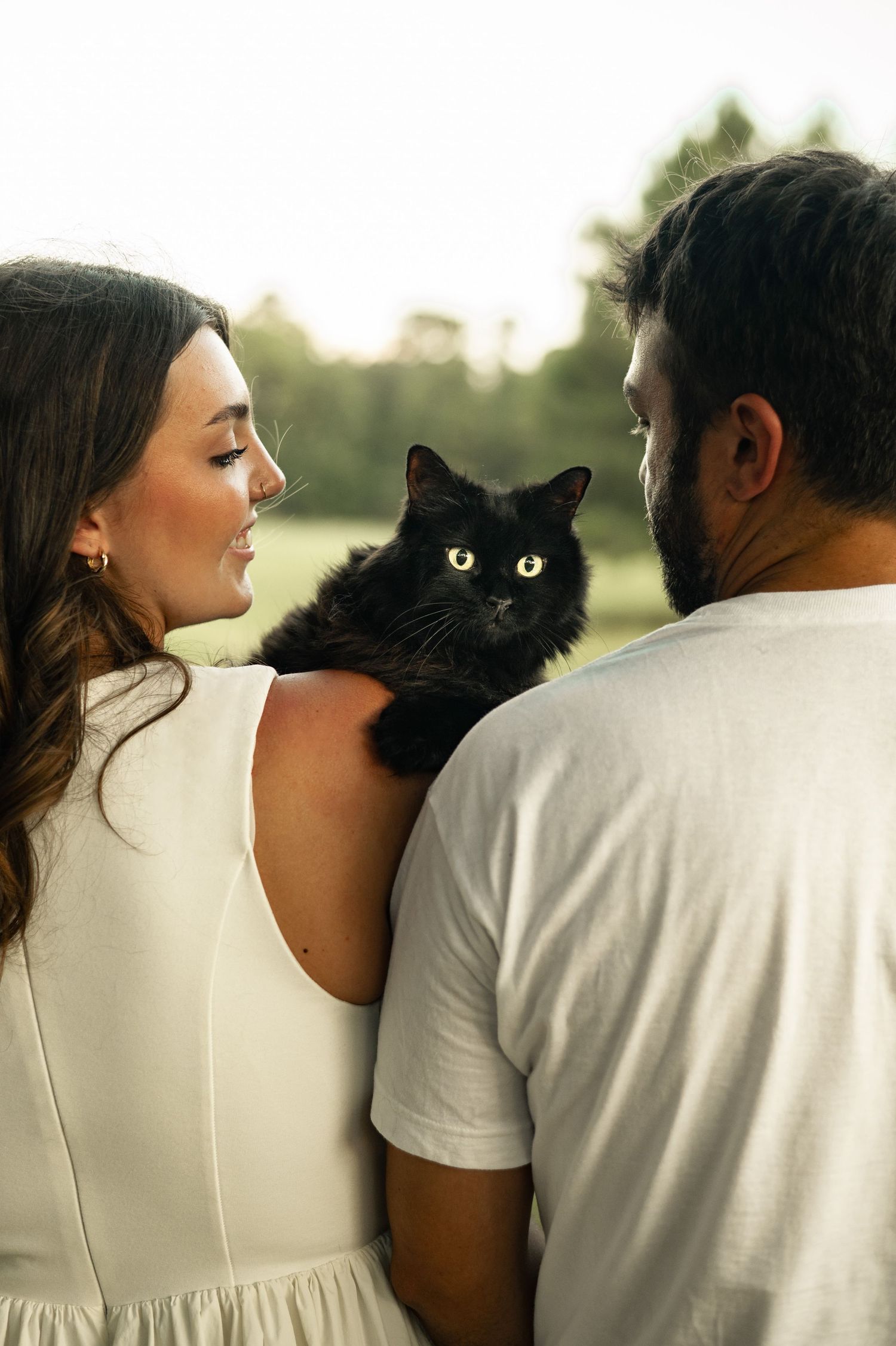 A couple in white clothing smiles at each other while holding a fluffy black cat outdoors.