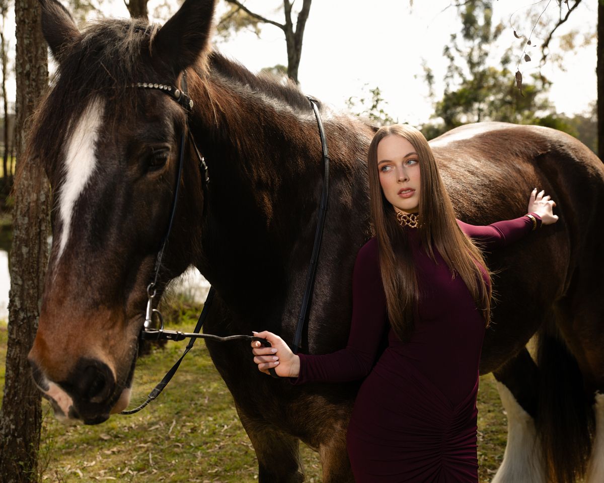 Young woman in burgundy dress poses with a dark brown horse in a forest setting.