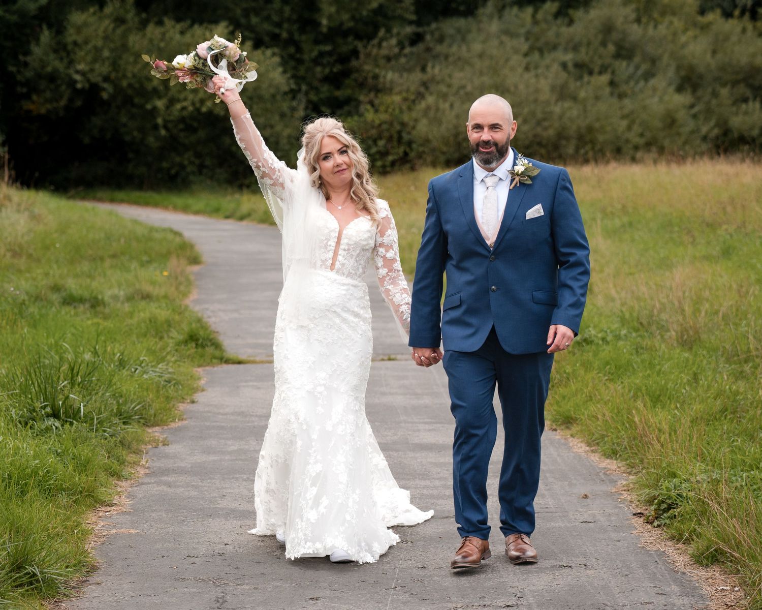 Bride in white lace gown raises bouquet while groom in navy suit holds her hand on a path.