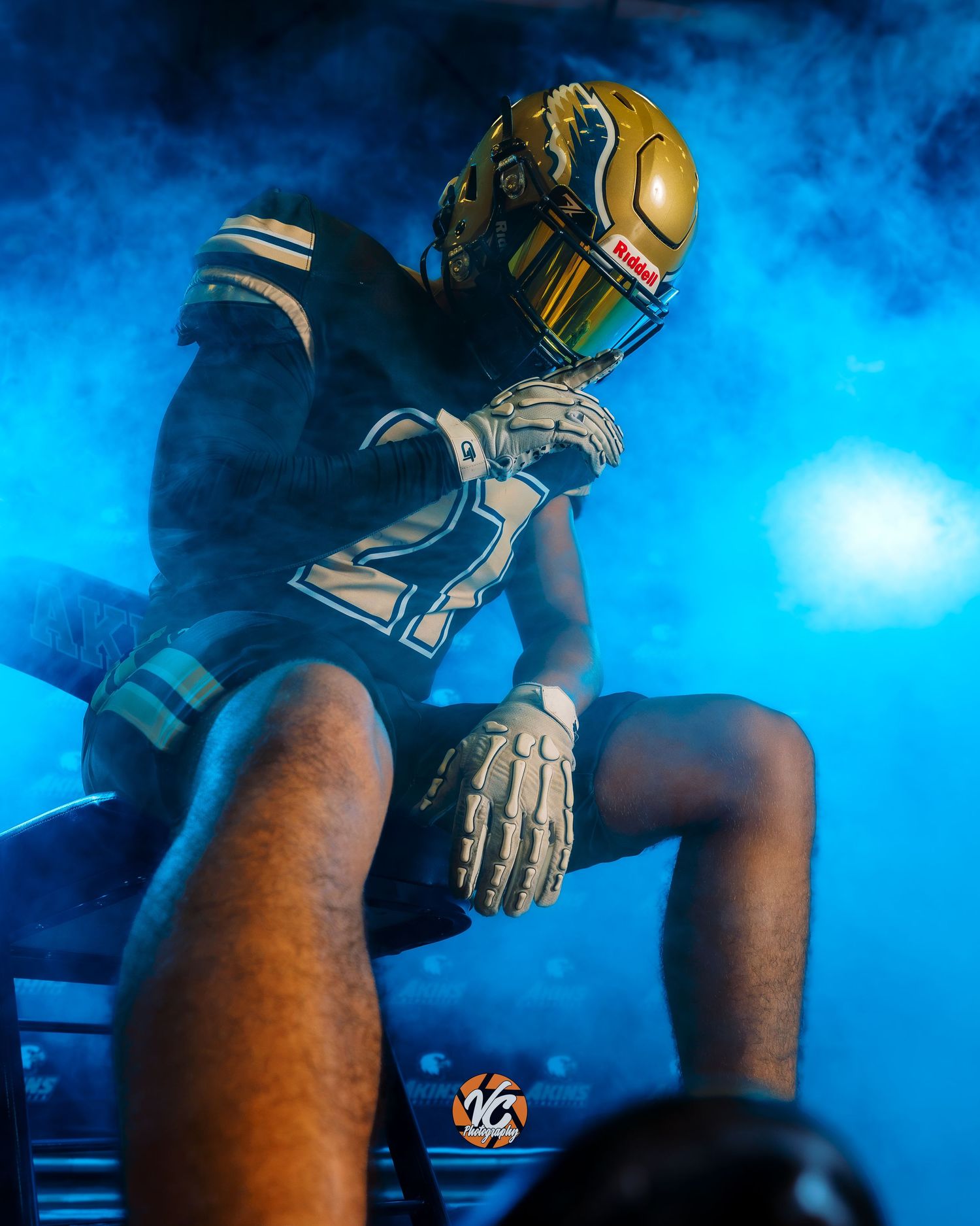 Football player in gold helmet and dark uniform poses dramatically in blue smoke during pre-game entrance.