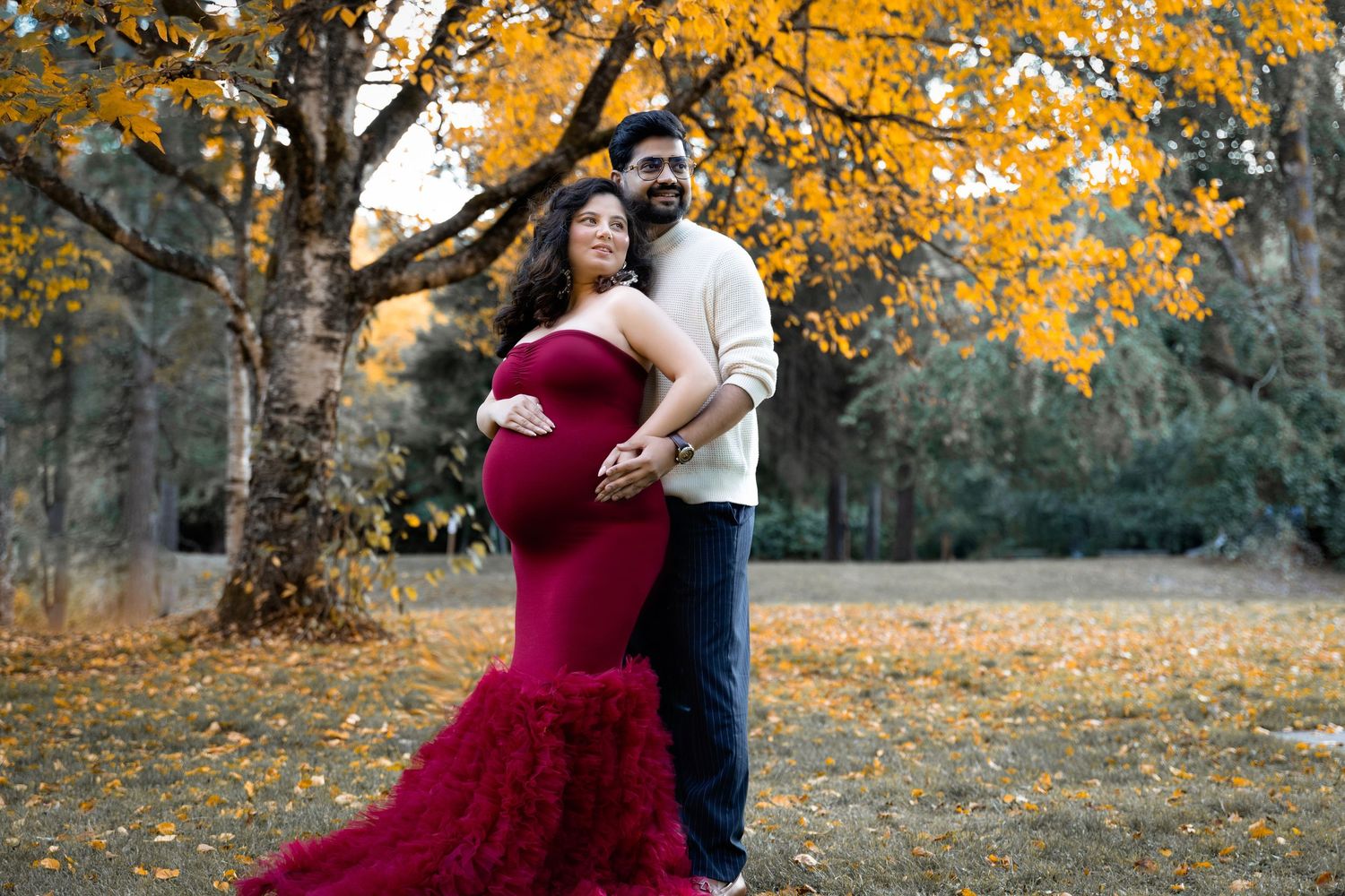 Couple poses for fall maternity photos under golden autumn tree, woman in red gown cradling baby bump.