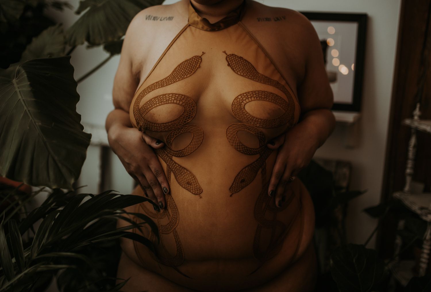 A moody boudoir photo highlighting the midsection of a curvy woman wearing snake embroidered lingerie and surrounded by lush plants