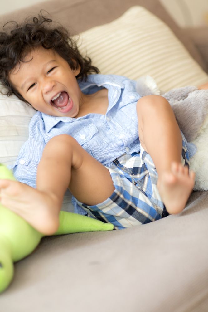 Adorable laughing toddler in blue shirt and plaid shorts playing on a beige couch with a green toy.