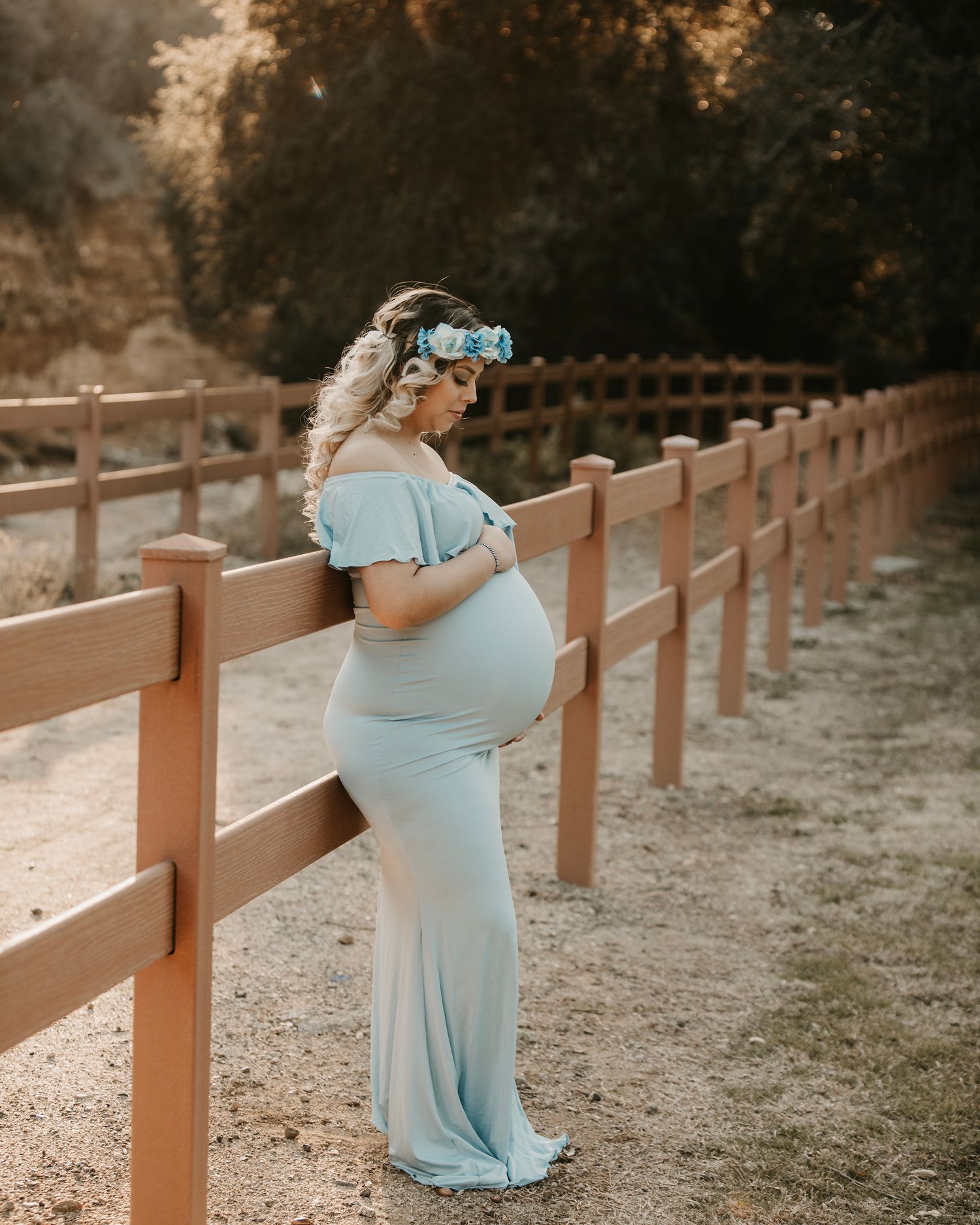 A light blue maternity dress photoshoot by a wooden fence at sunset.