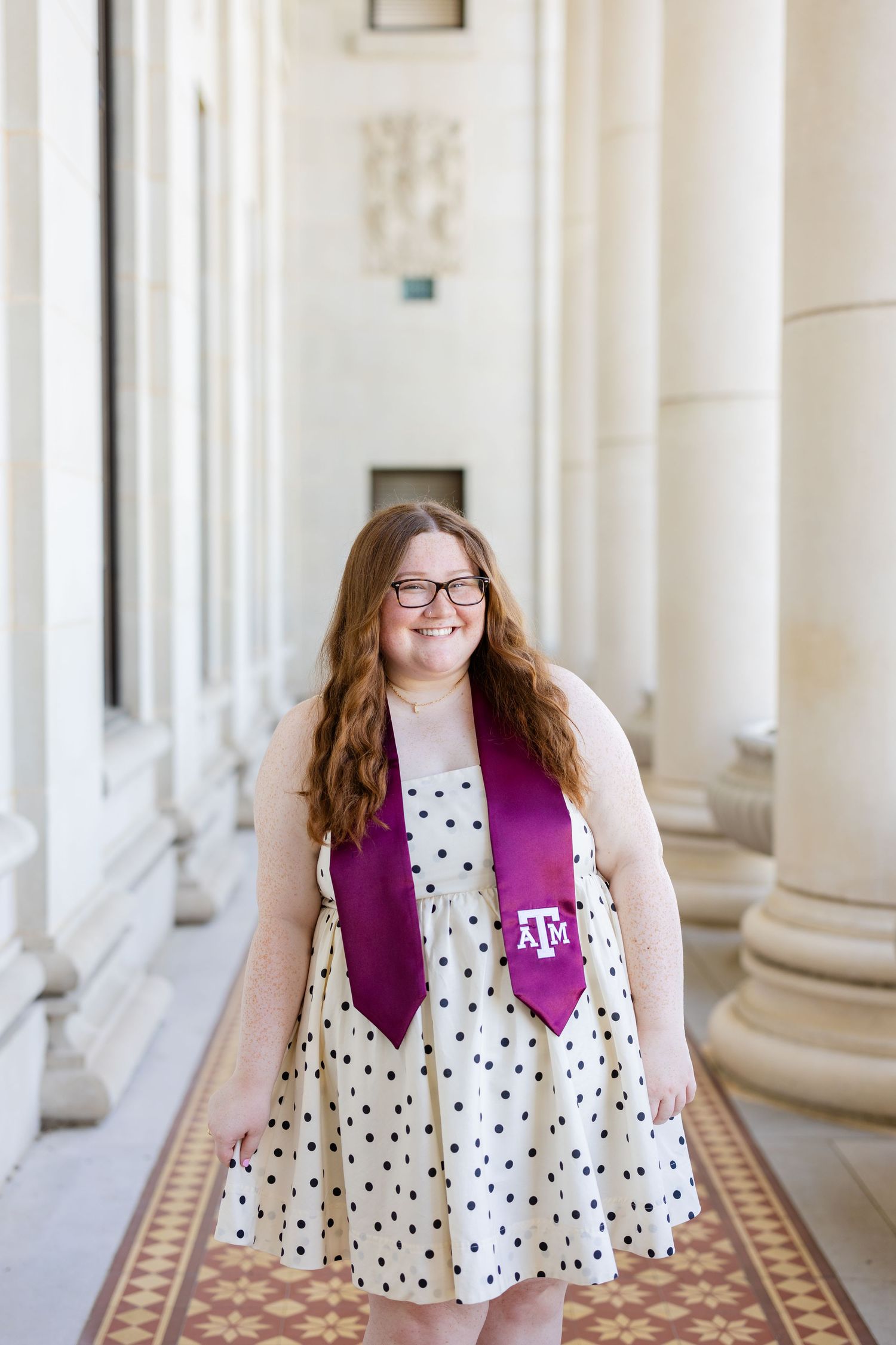 Graduate in polka dot dress and maroon Texas A&M graduation stole poses between white columns.