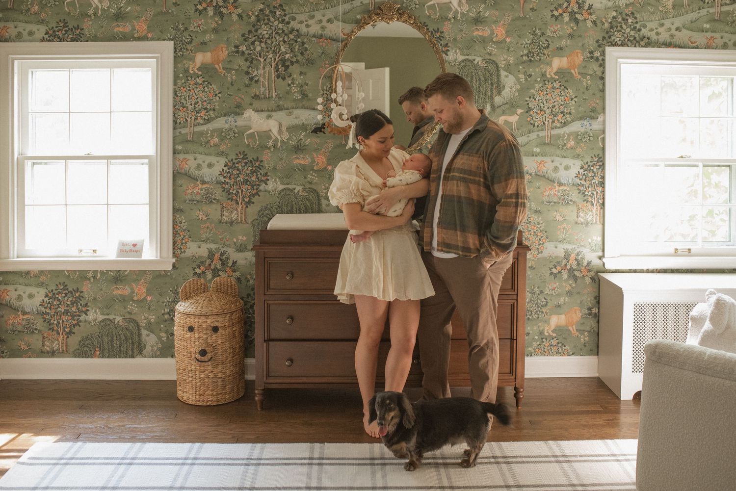 Couple standing together in vintage-style room with floral wallpaper, wooden dresser, and black dog on hardwood floor.