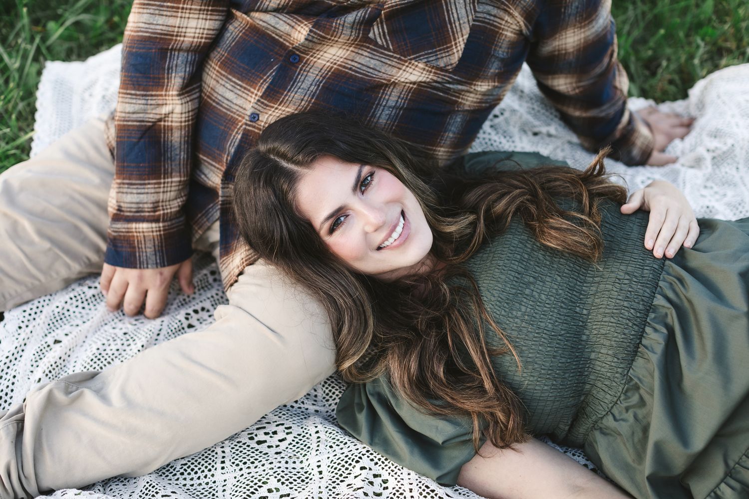 Woman with long dark hair wears eucalyptus dress lays on fiancee's lap wearing tan pants and plaid flannel for engagement photos in Concord.
