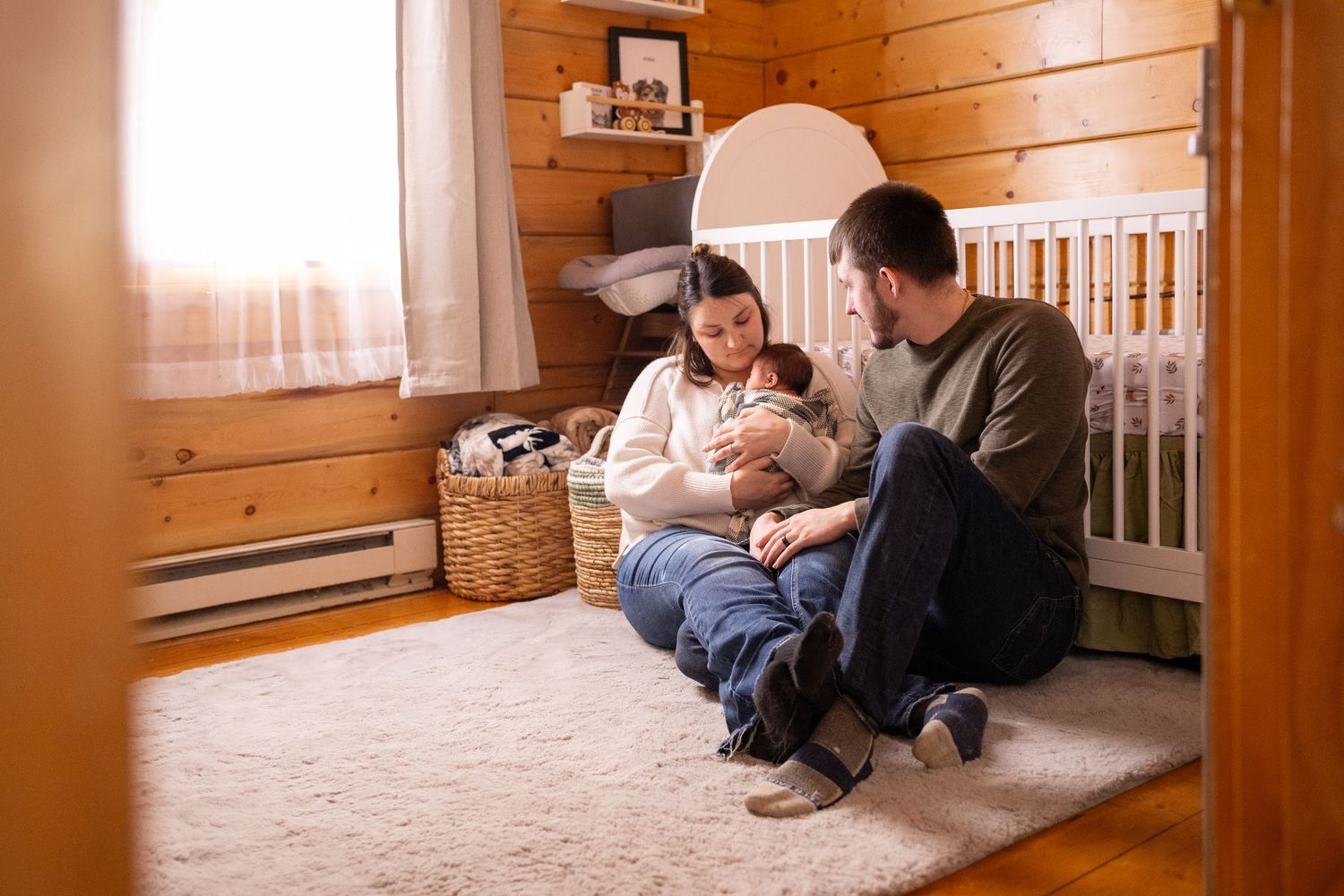 A cozy wood-paneled nursery room with a crib and people sitting on the floor.
