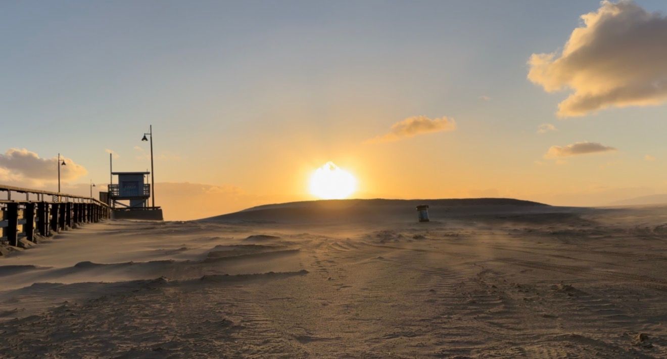 A panoramic sunset view of a sandy beach with rolling dunes and wooden boardwalk stretching into the distance.