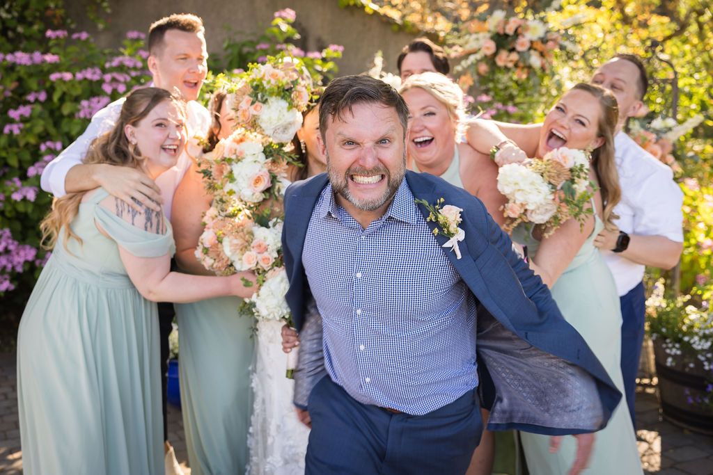 Groom grimaces playfully as bridal party celebrates behind him at outdoor wedding with floral backdrop.