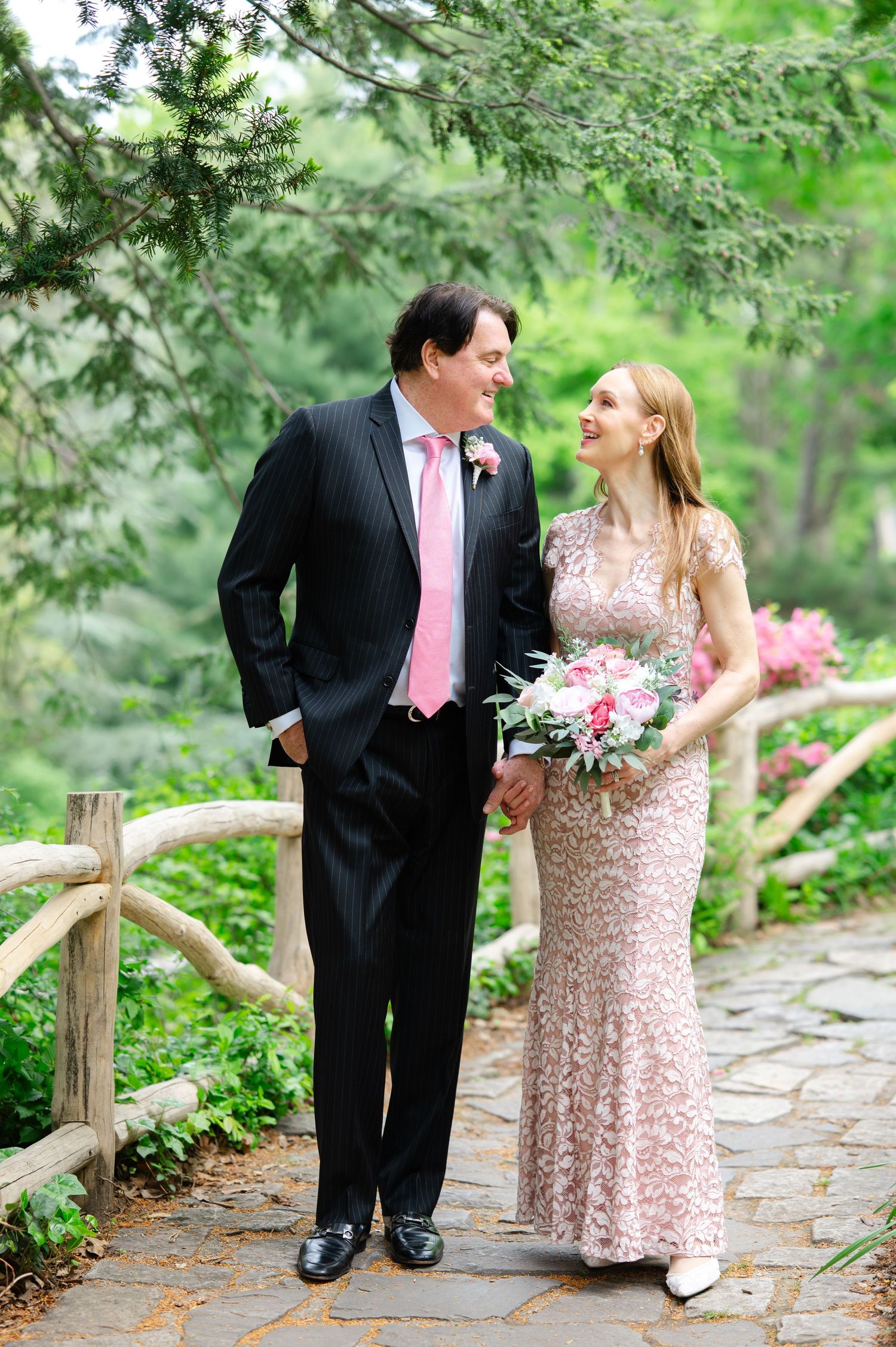 A couple in formal attire walks hand-in-hand on a stone path surrounded by lush greenery.