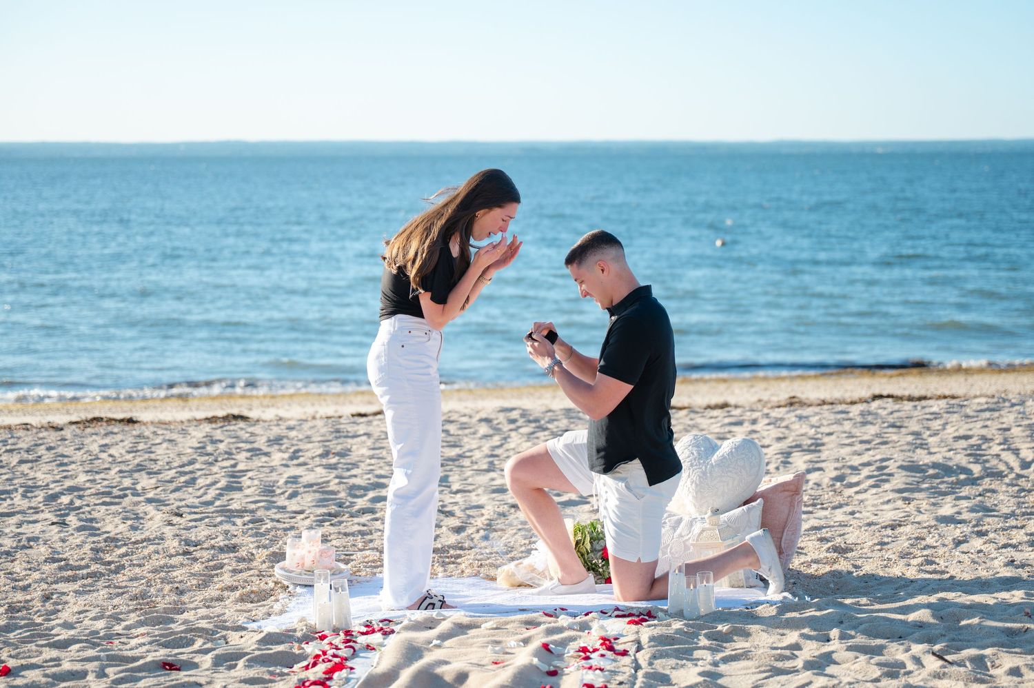 Man proposes to woman on sandy beach with ocean backdrop, rose petals scattered around them.