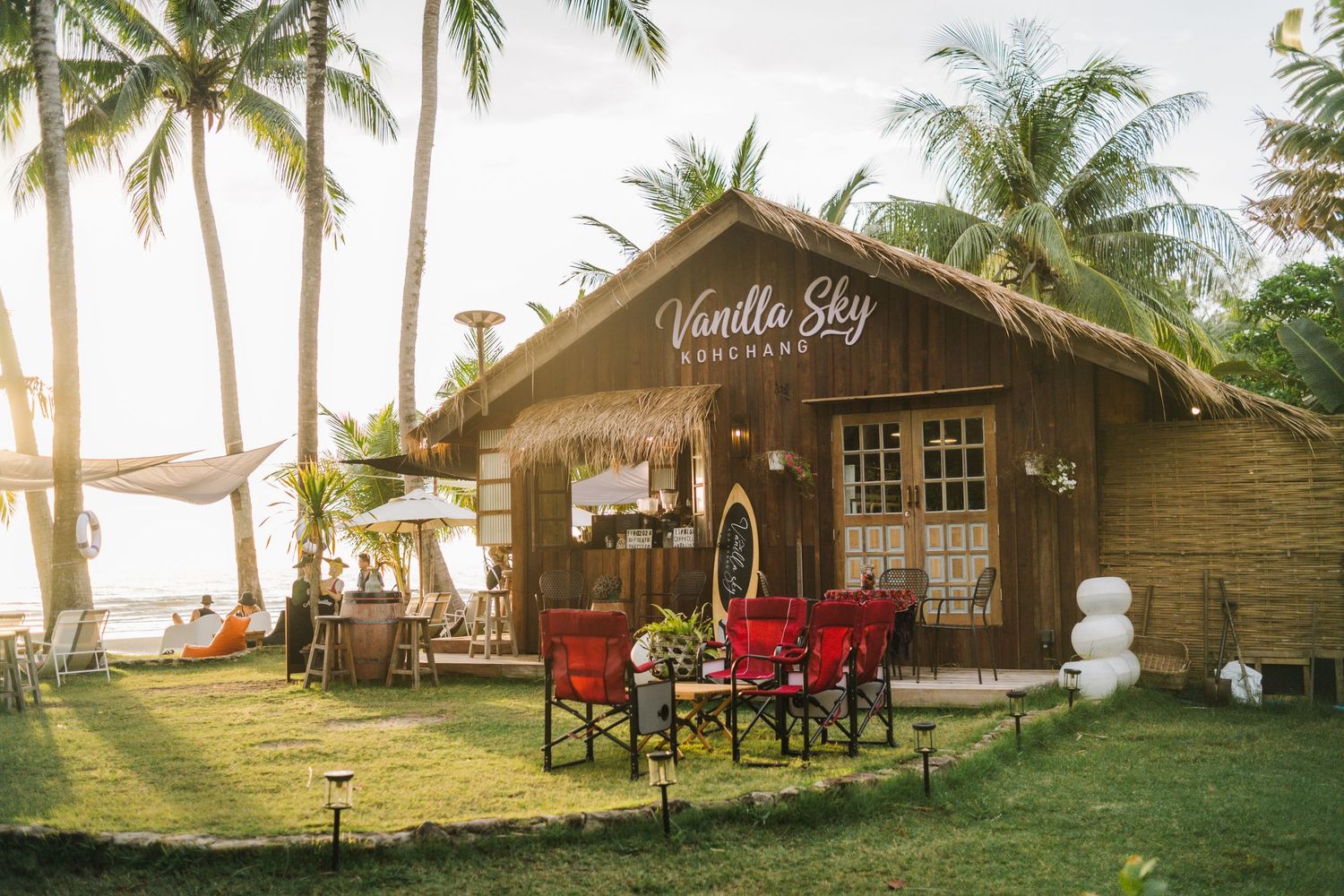 Vanilla Sky beachside restaurant with wooden cabin, red chairs, lush palm trees, and ocean view at golden hour.