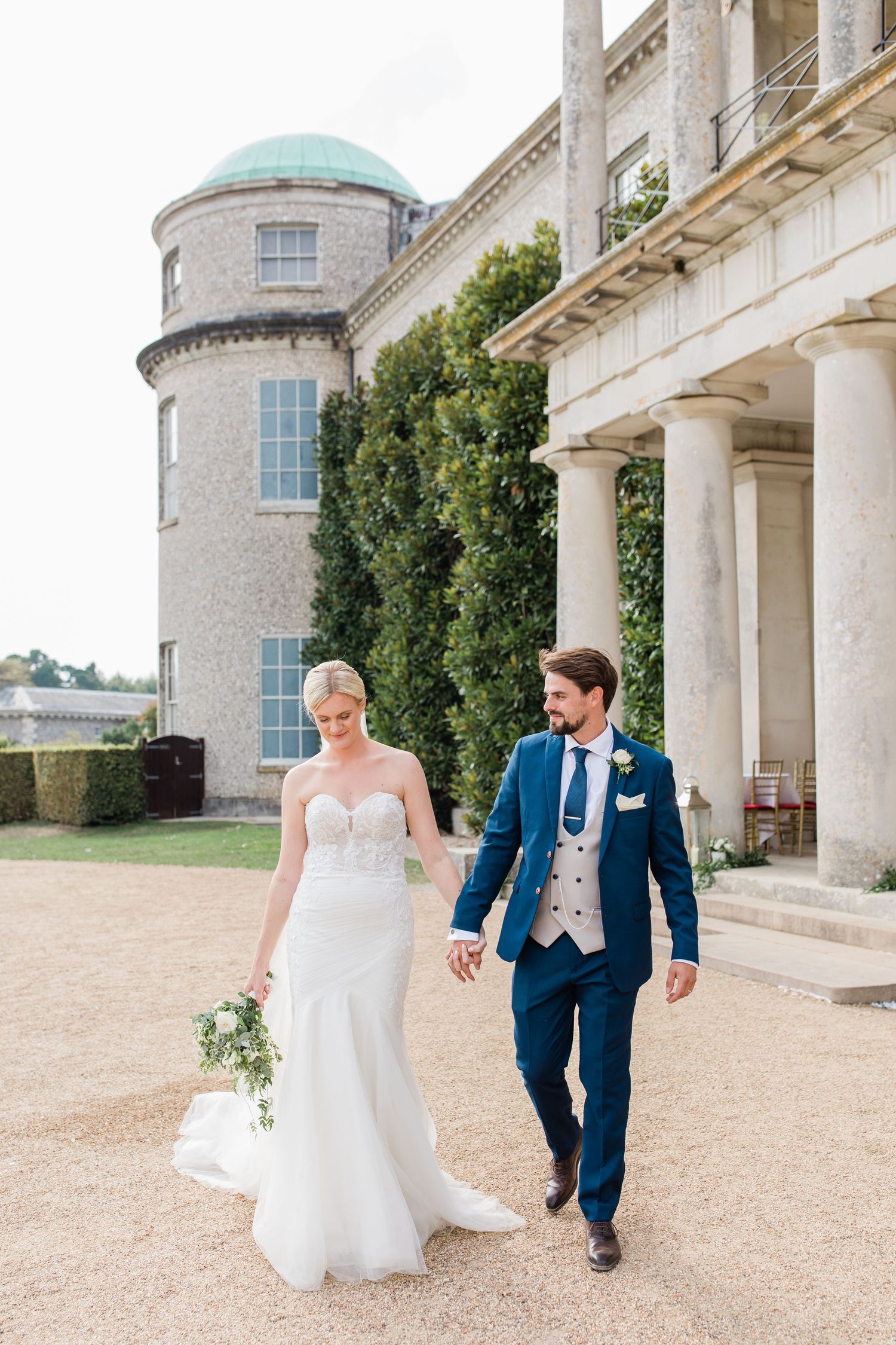 A couple walks together outside a grand historic mansion with stone columns and turret on a sunny day.