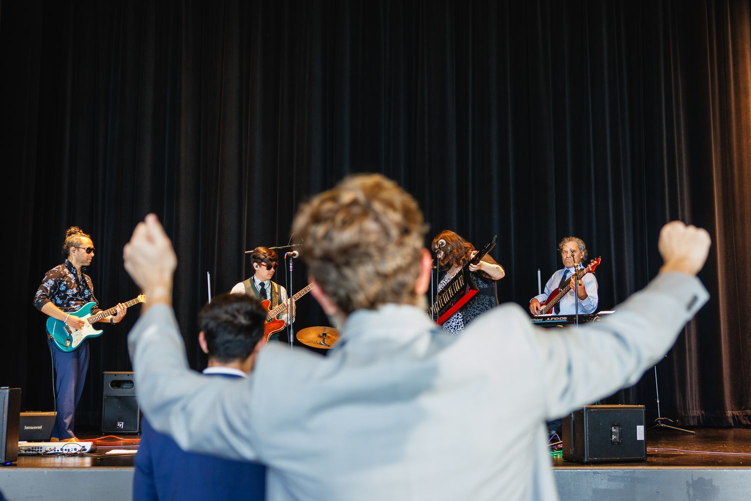 Audience member raises fists while a live band performs on stage with dark curtain backdrop.