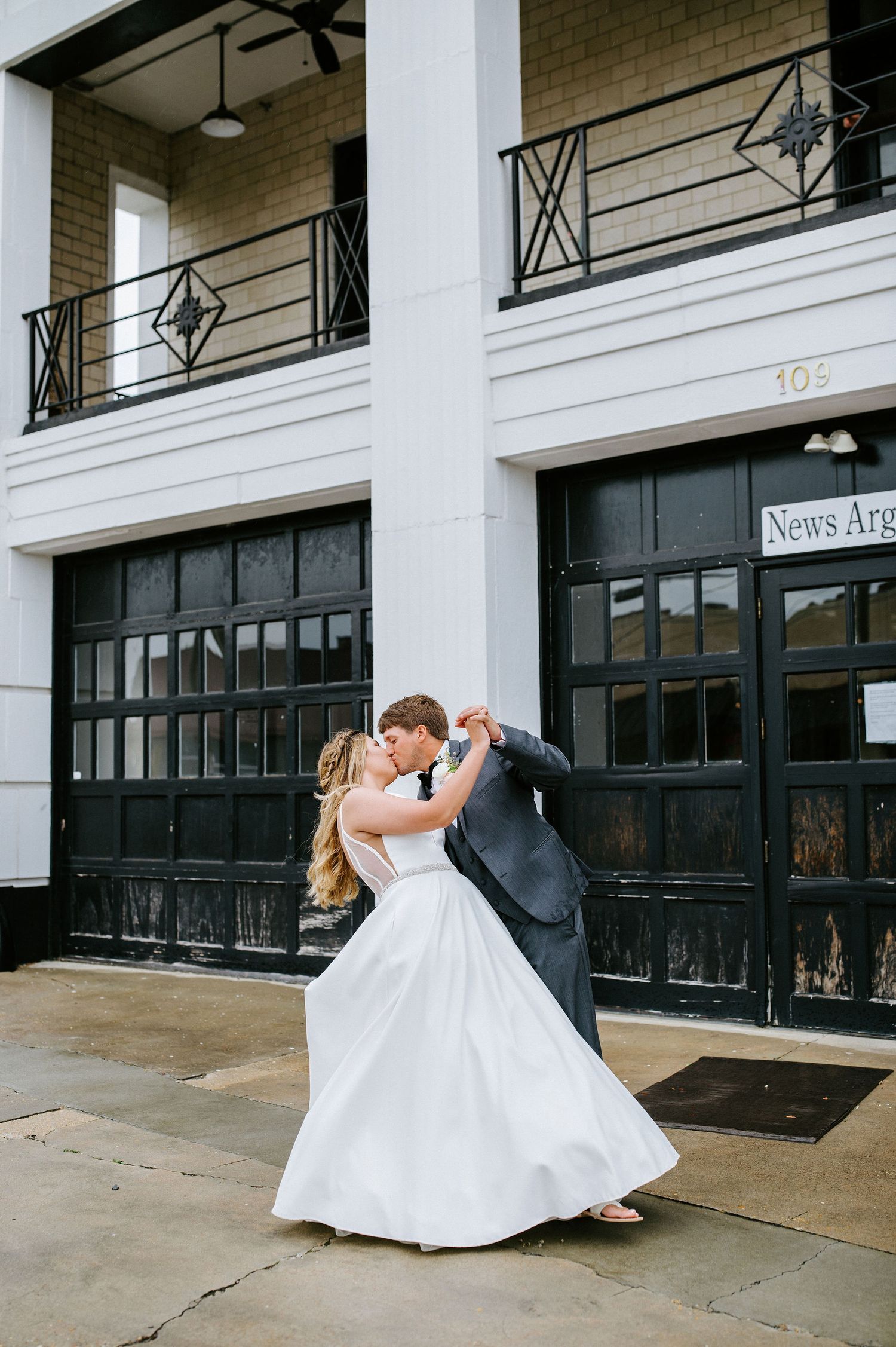 A romantic wedding couple dips and kisses in front of a brick building with black storefront windows.
