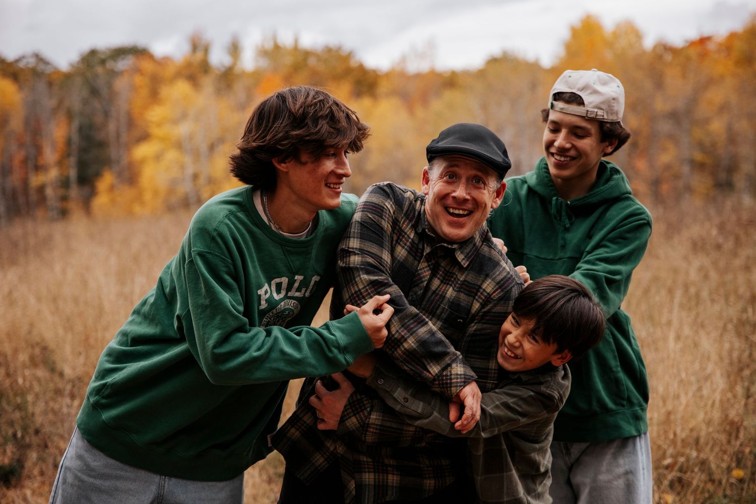 Three people wearing green and plaid shirts stand together in a field with autumn foliage in the background.