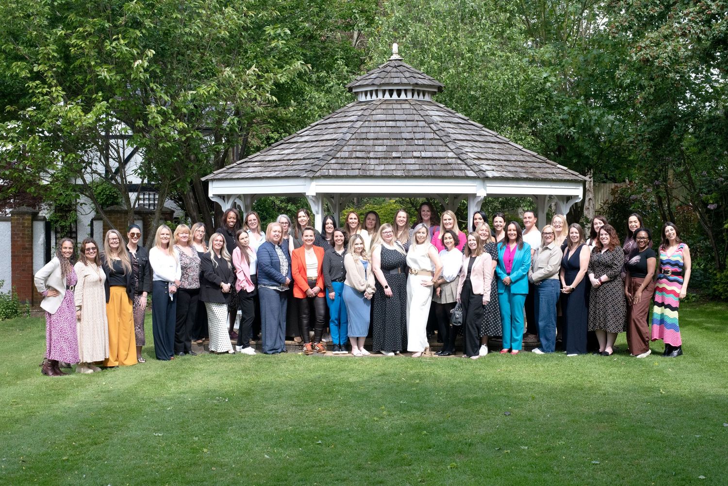 Large group of elegantly dressed people posing outdoors near a white gazebo surrounded by lush green trees.