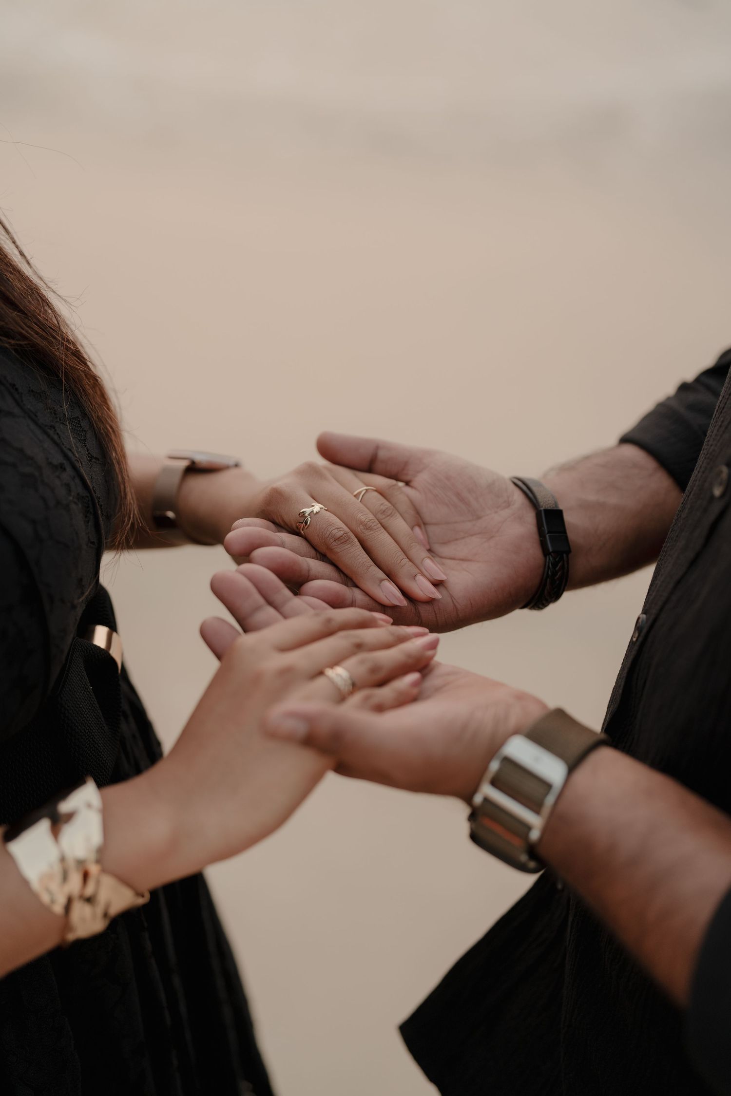 Couple holding hands on beach, wearing gold rings, bracelets, and watches, dressed in black outfits.