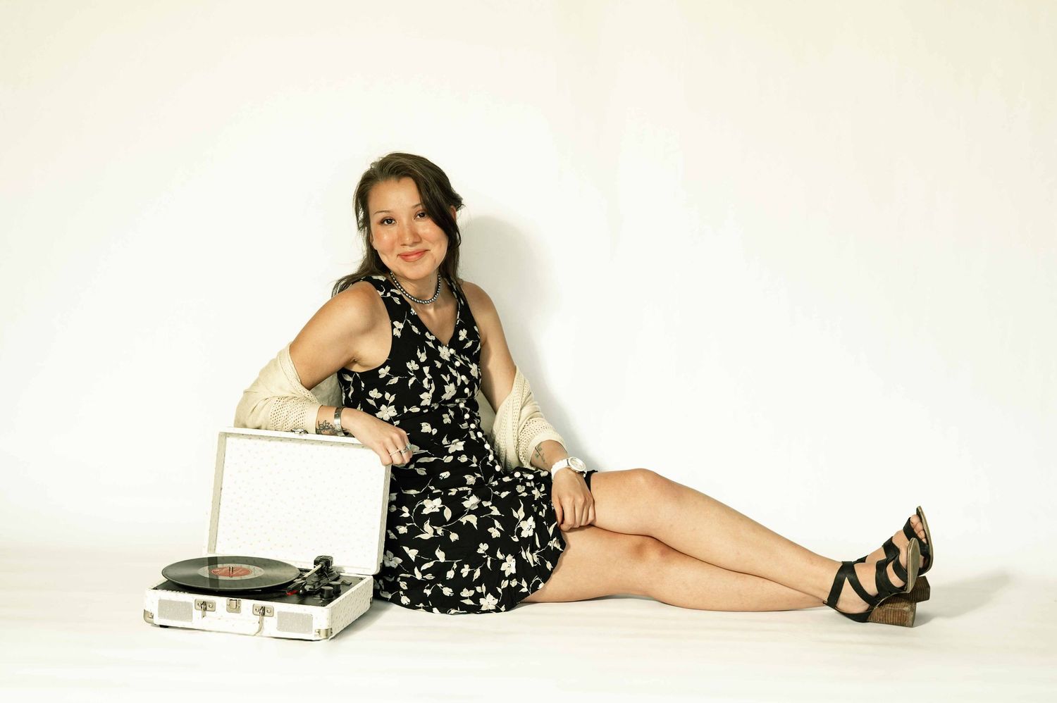 Young woman in floral dress sitting on white floor beside vintage record player turntable, smiling at camera.