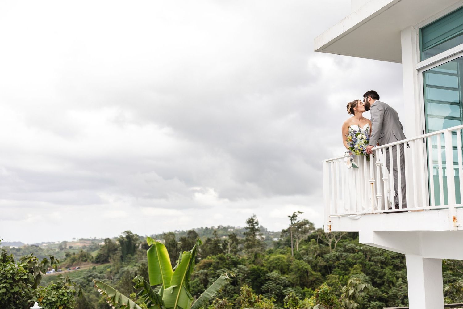Bride and groom kissing on a white balcony overlooking lush tropical greenery under a cloudy sky.