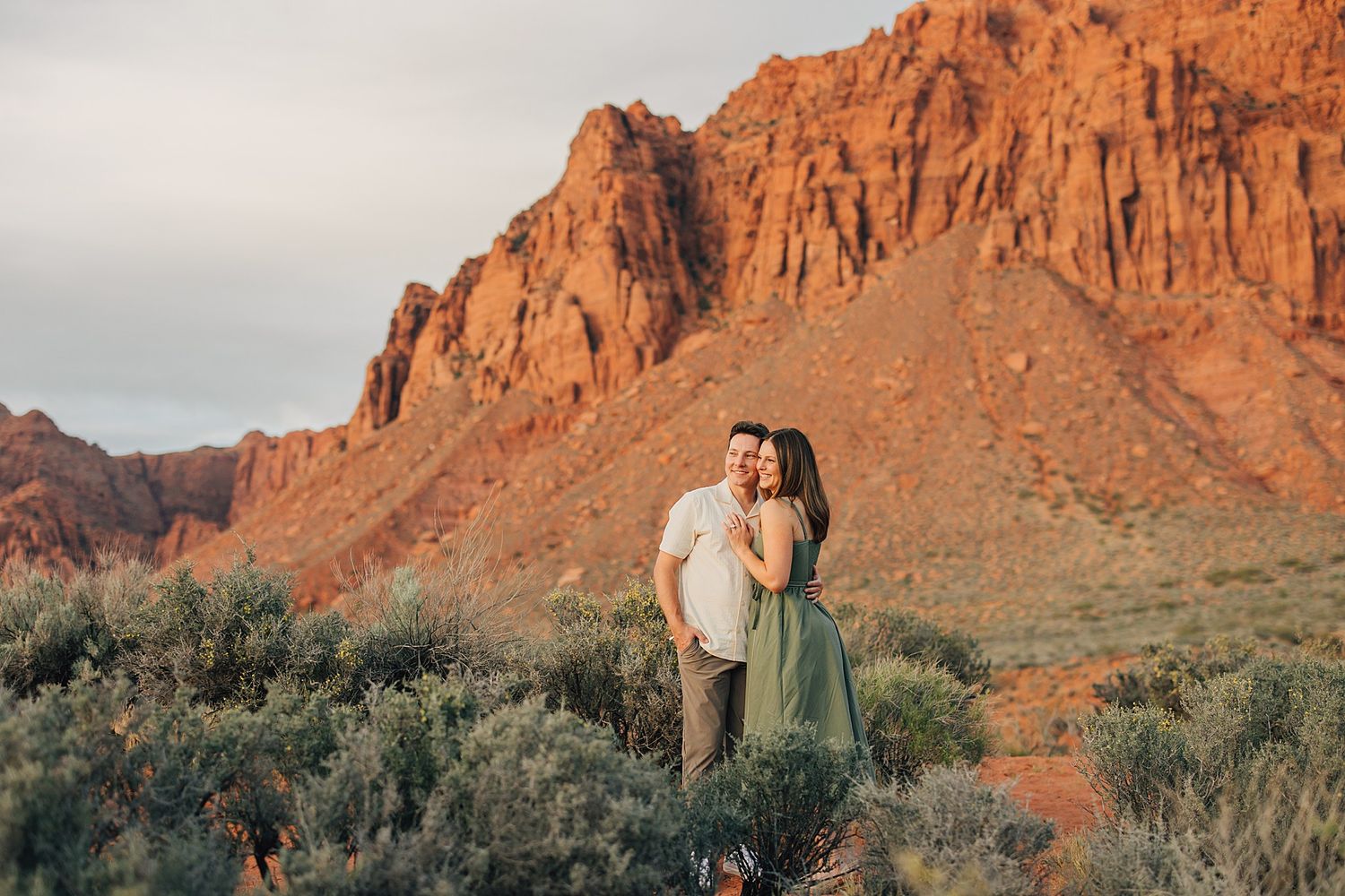 Couple embracing in front of red rock cliffs in desert landscape at golden hour with green sagebrush.