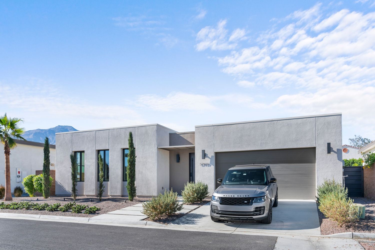 Modern single-story stucco home with flat roof, two-car garage, desert landscaping, and mountain views under blue sky.