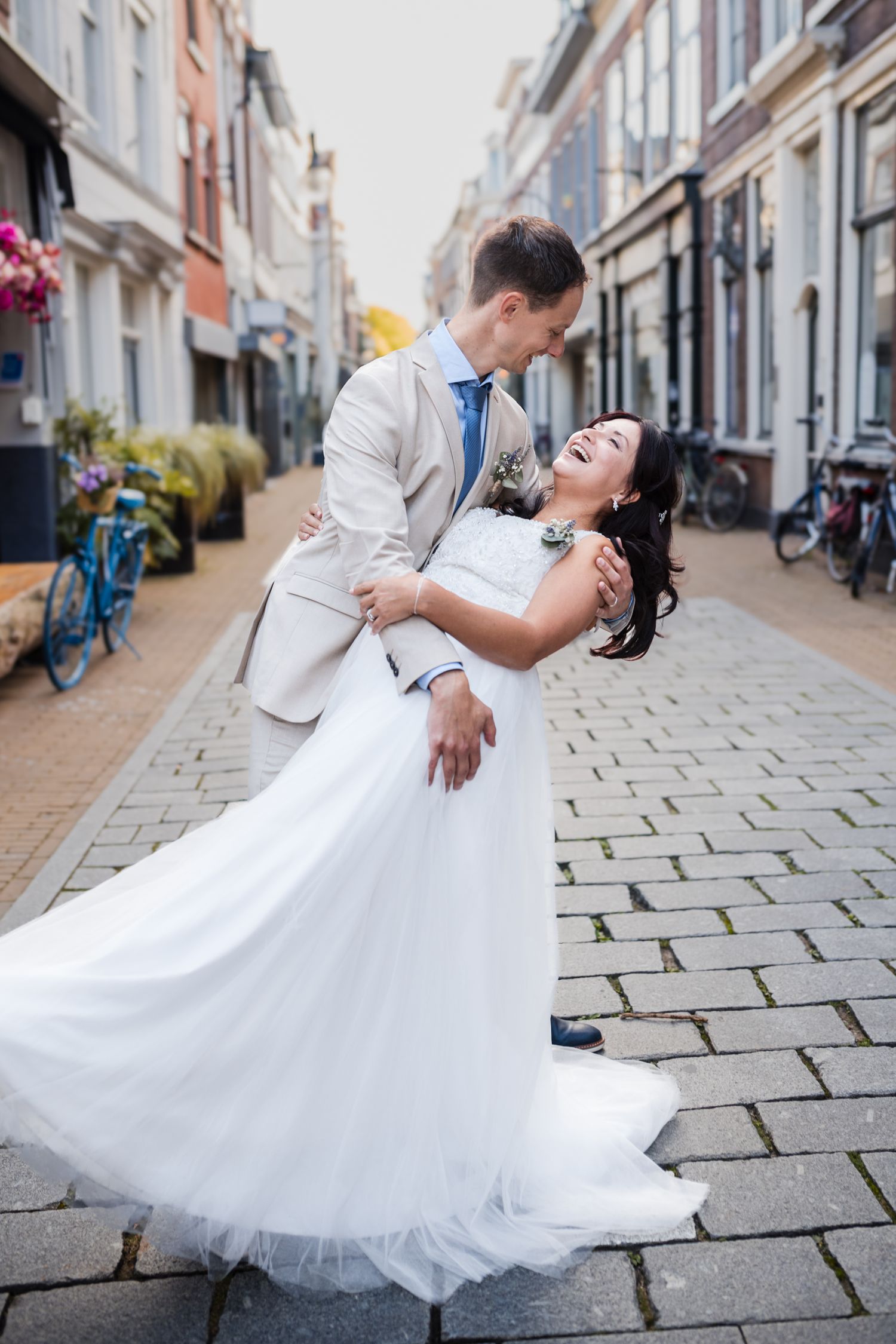 A joyful bride in a white gown and groom in a beige suit share a romantic dip on a cobblestone street in Gorinchem.