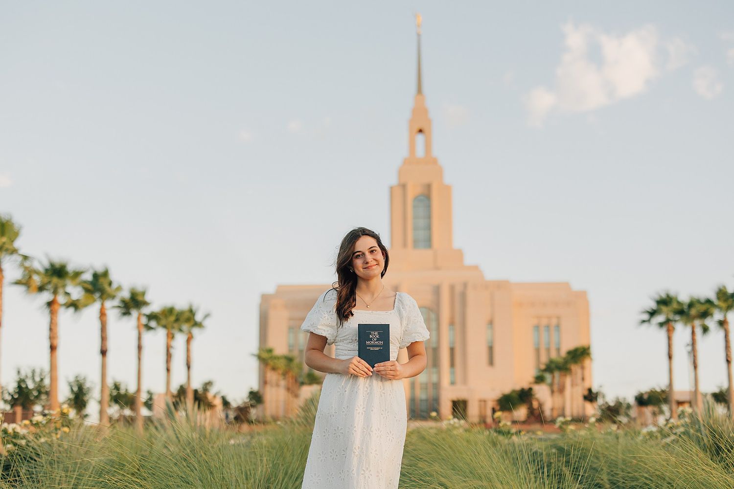 Young woman in white dress holding a book outside an LDS temple surrounded by palm trees.