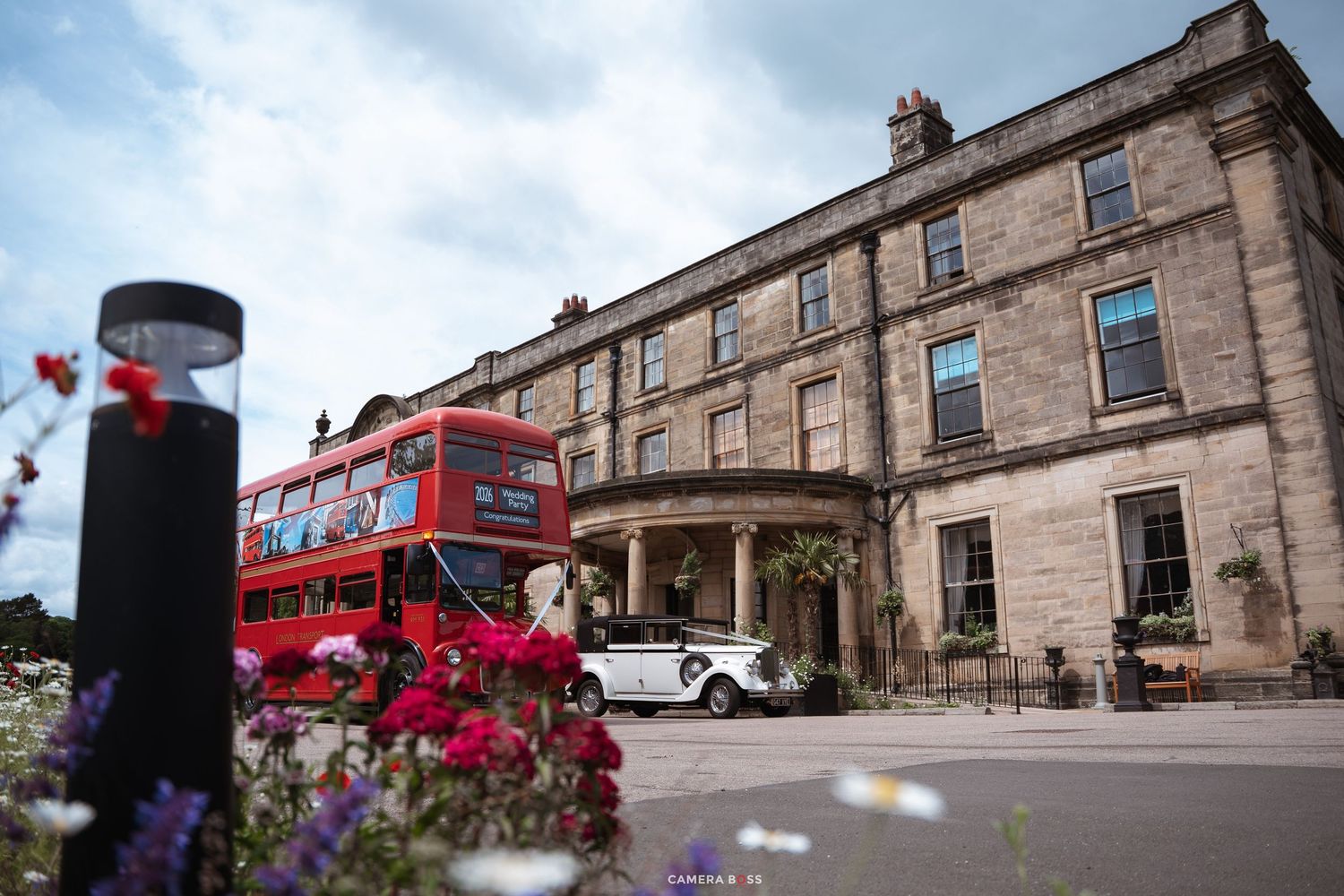 A red double-decker bus and white vintage car outside a grand stone manor house with colorful flowers in the foreground.