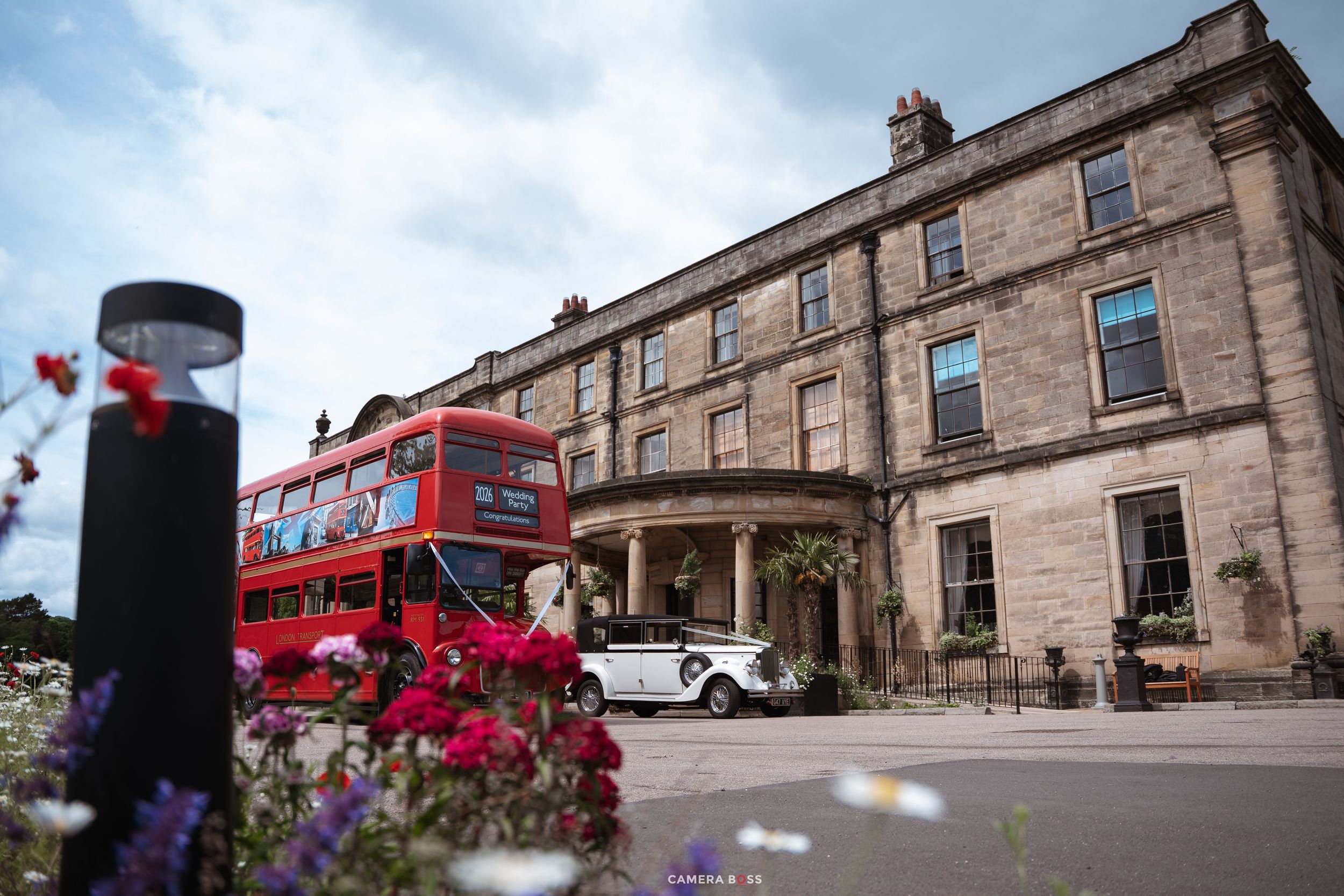 A red double-decker bus and white vintage car outside a grand stone manor house with colorful flowers in the foreground.
