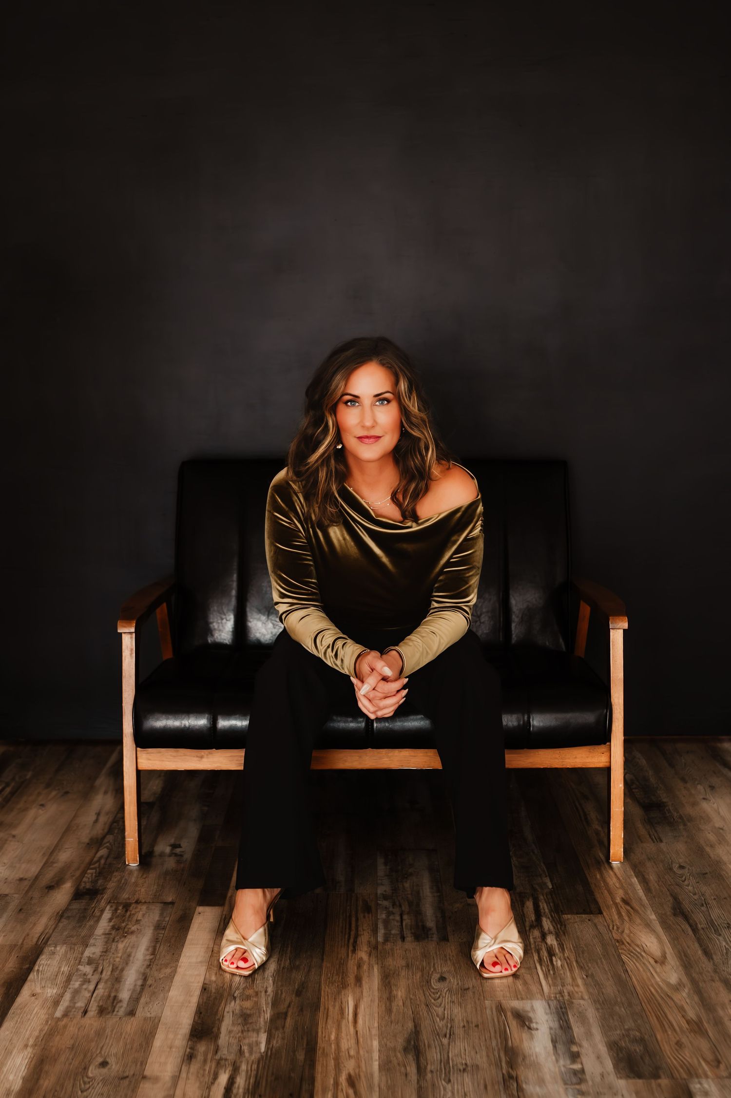 Confident woman in olive velvet top seated on black leather chair against dark studio background.