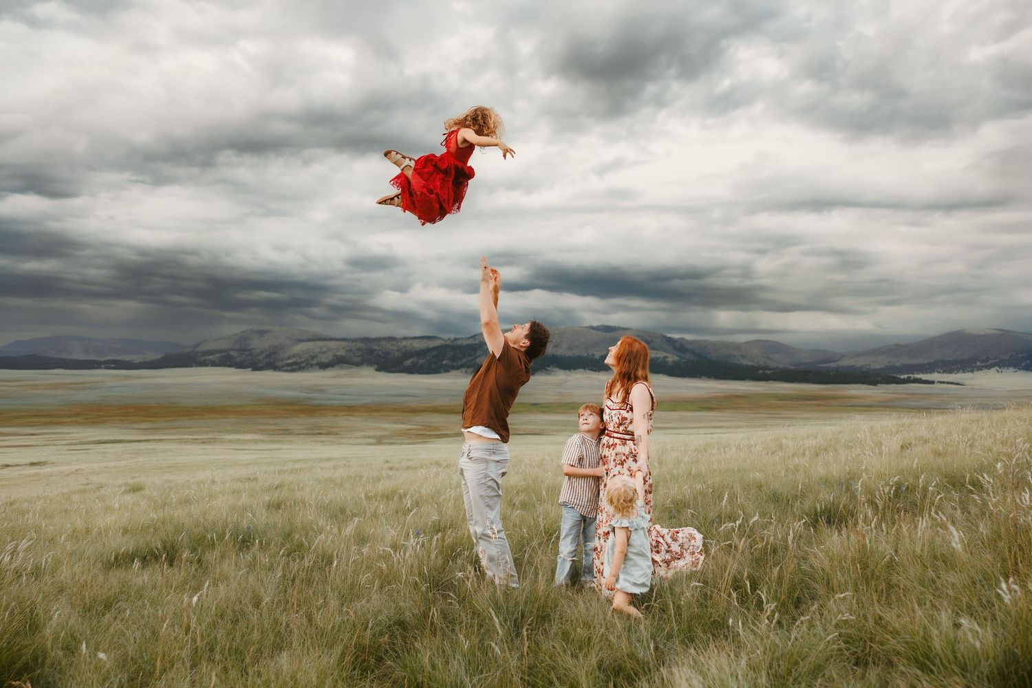Family in a field tossing a child in a red dress into the air under dramatic stormy skies.