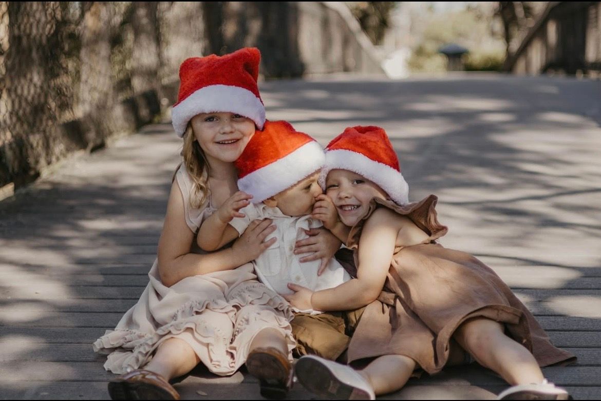 Three young girls wearing Santa hats sit together on a wooden bridge, smiling and hugging during Christmas.