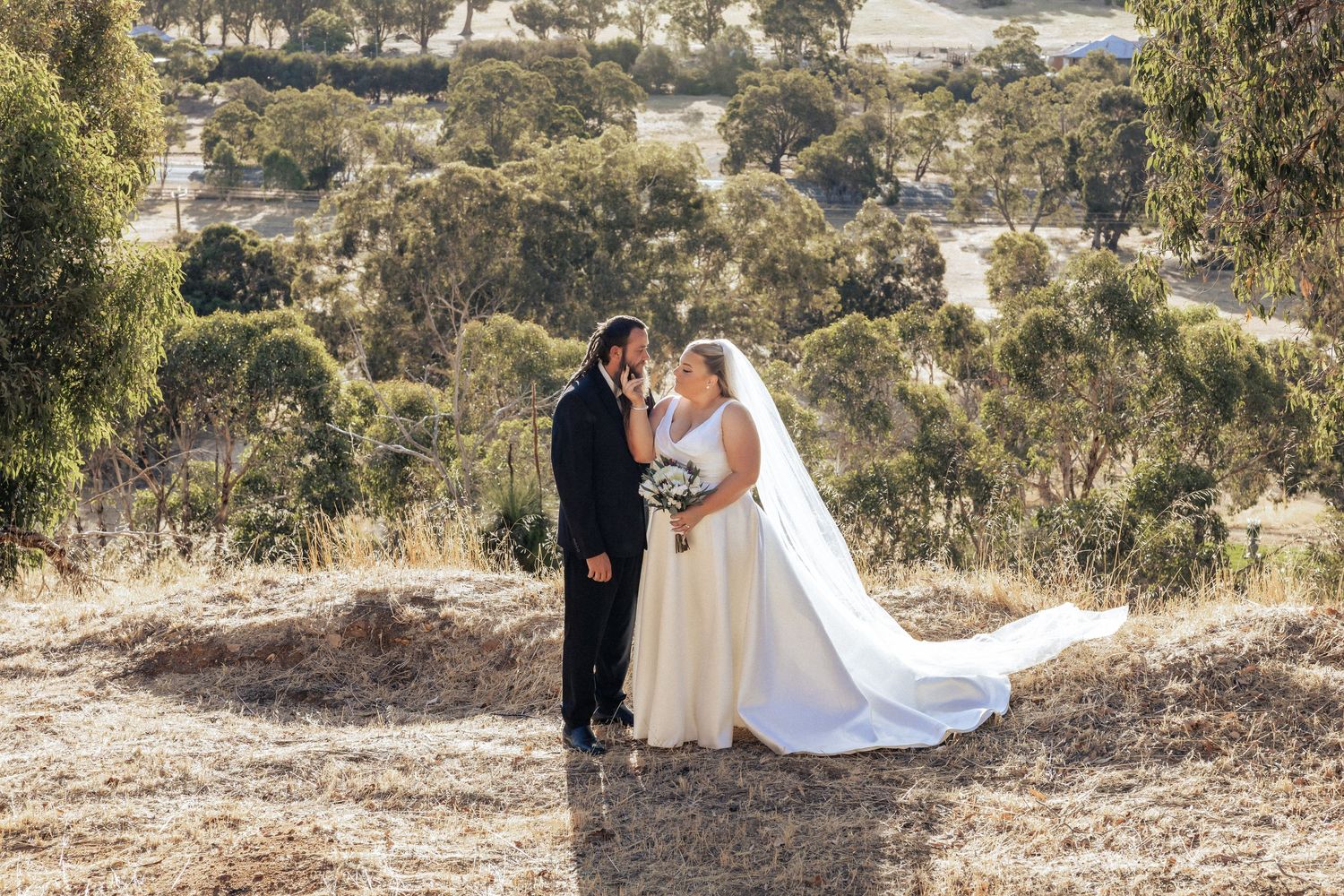 Bride and groom share a romantic kiss on a hilltop with scenic Australian countryside and eucalyptus trees in the background.