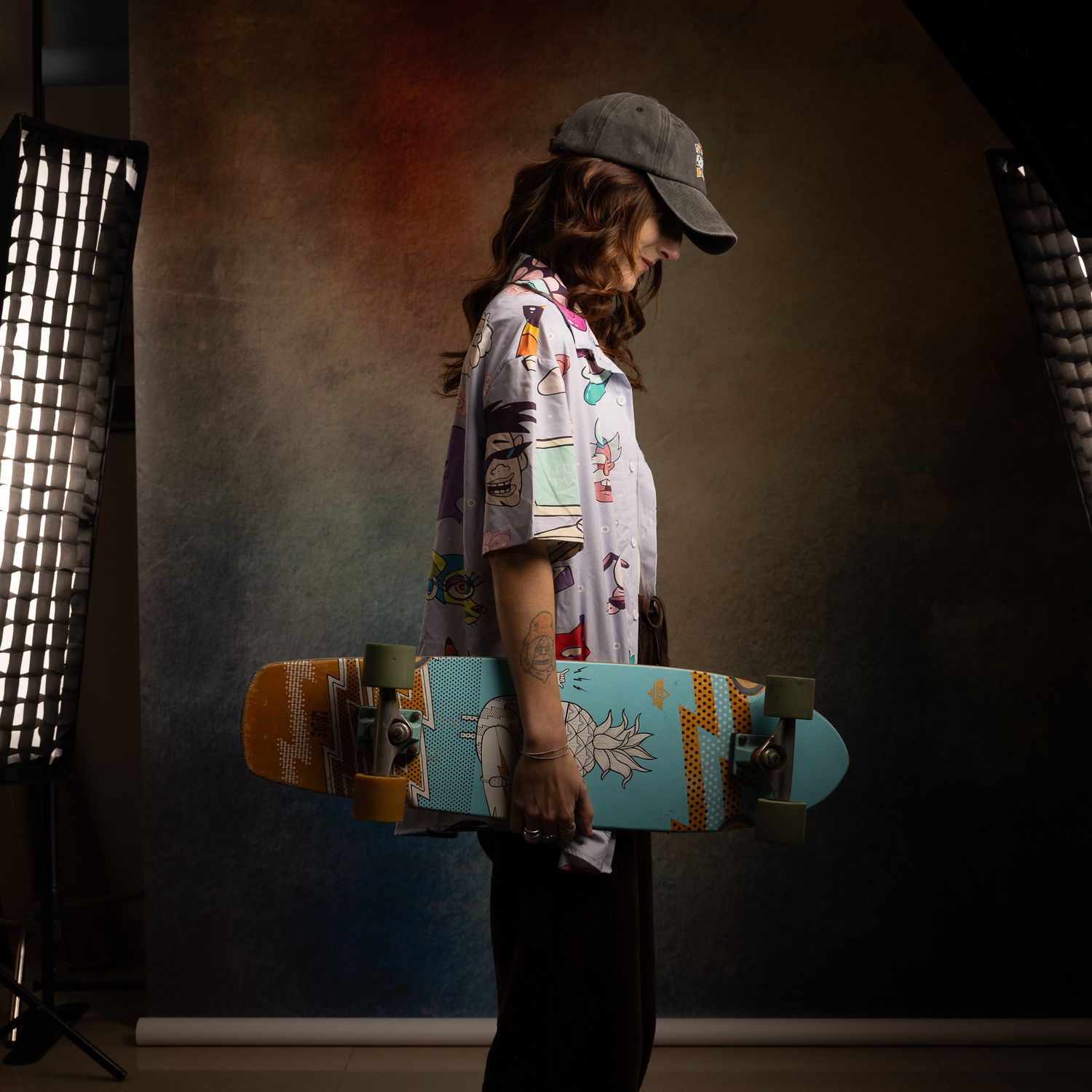A young person in a floral shirt and cap holds a teal skateboard, posing in front of a studio backdrop.