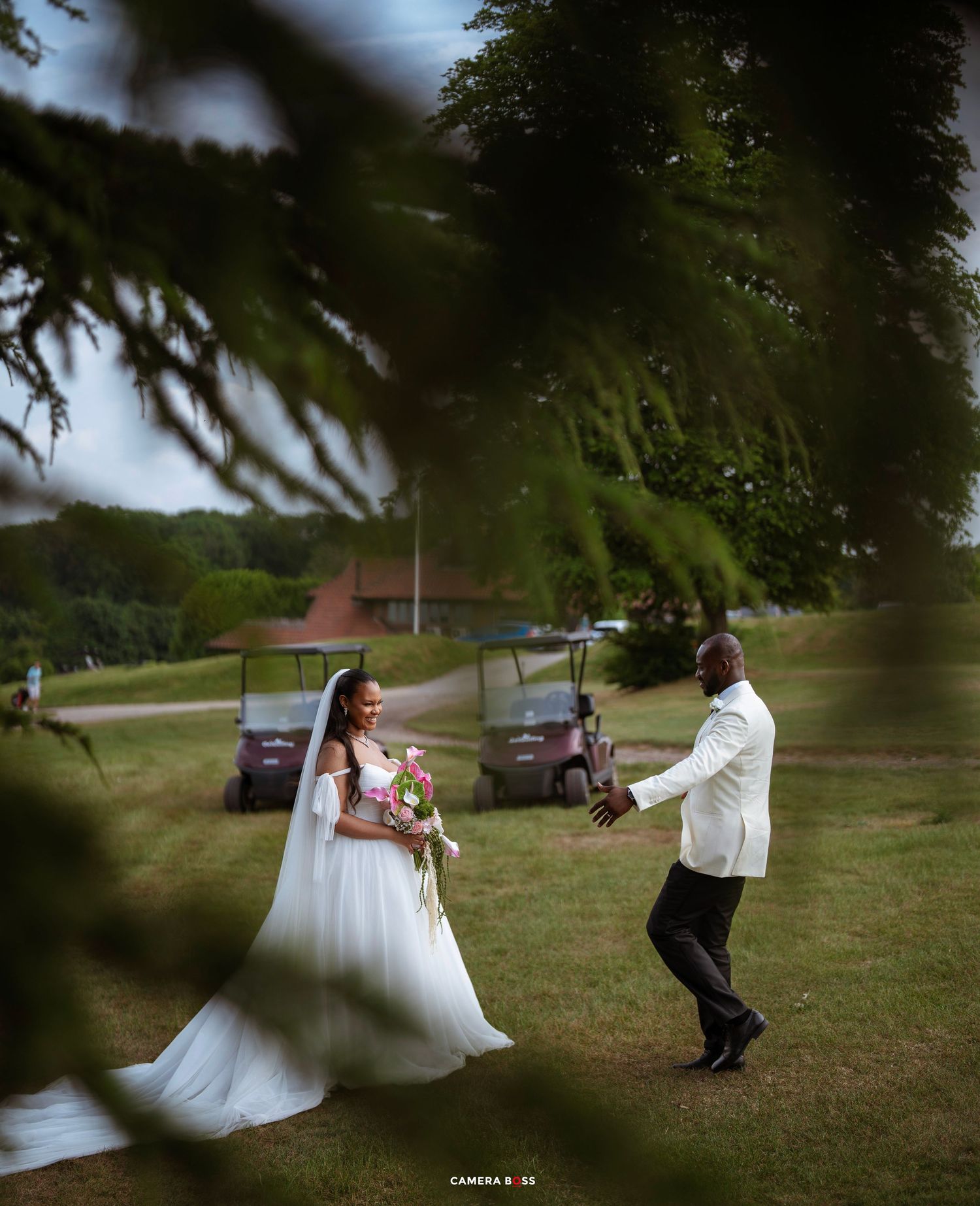 Bride in white gown holds bouquet as groom in white jacket reaches out on a lush green golf course.