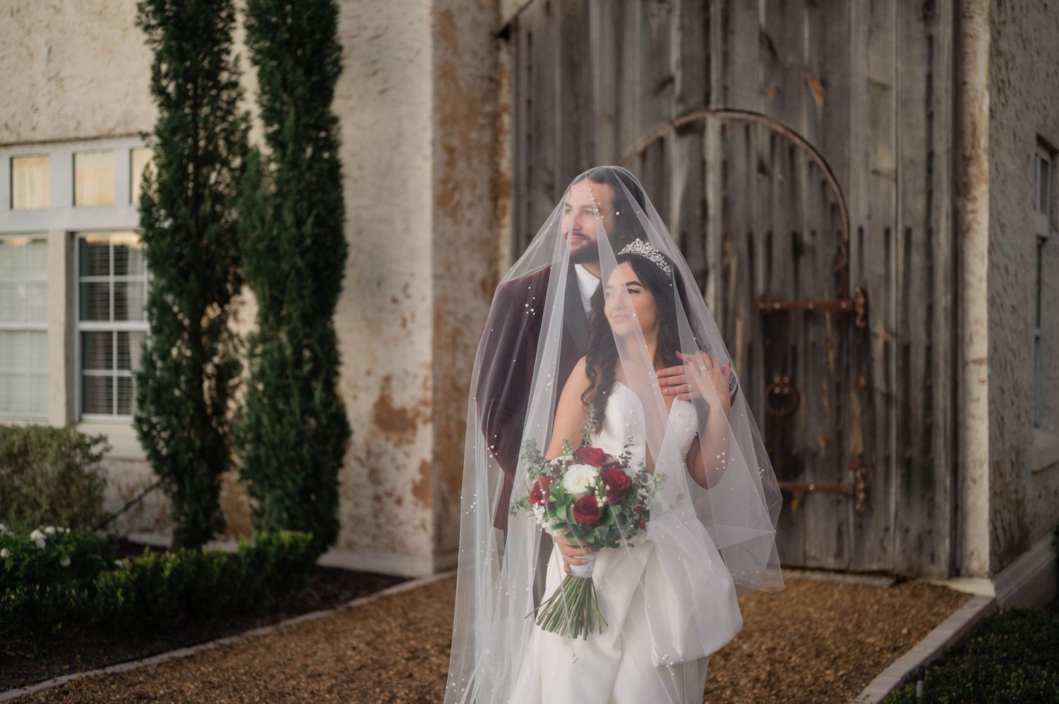 Bride in white wedding dress holds bouquet while walking down rustic barn pathway with wooden walls and greenery.