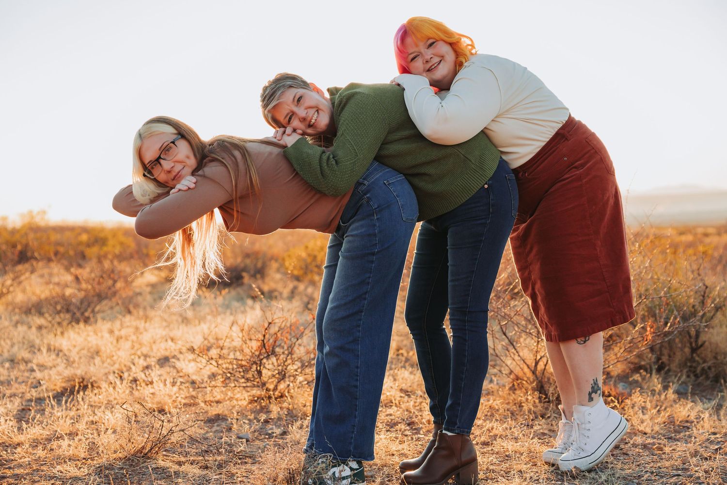 Three friends laugh together while posing playfully outdoors in a golden field at sunset.