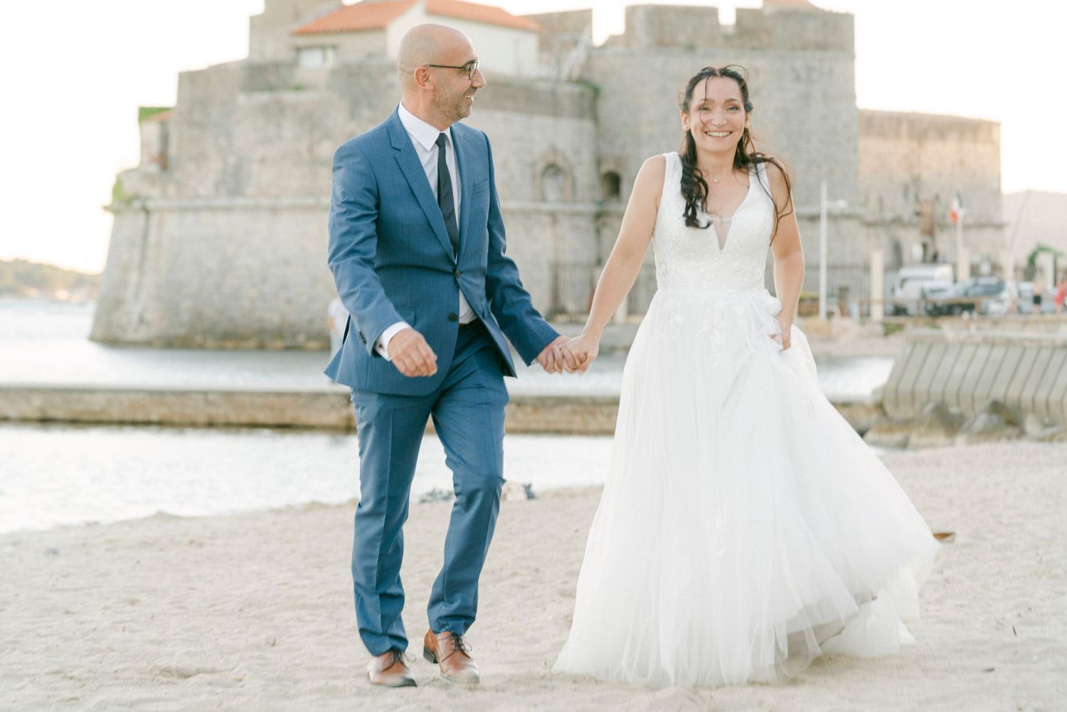 A couple in wedding attire walks hand in hand through a scenic vineyard on a sunny afternoon.