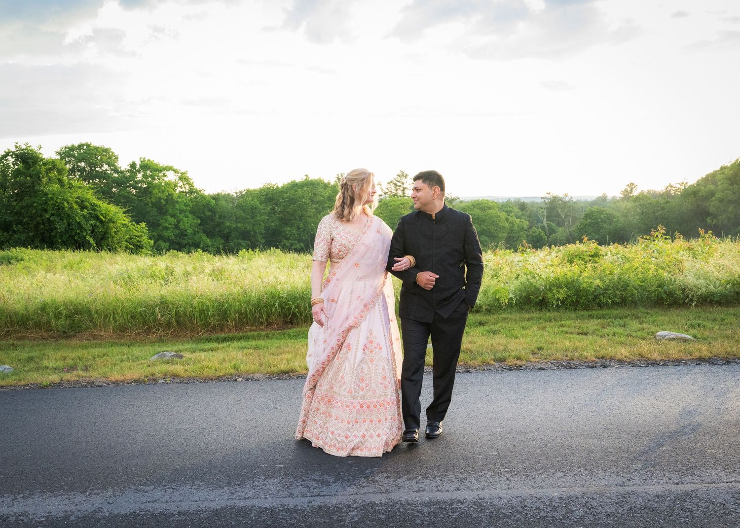Bride in white gown and groom in black tuxedo posing on a paved path with lush green fields and trees behind them.