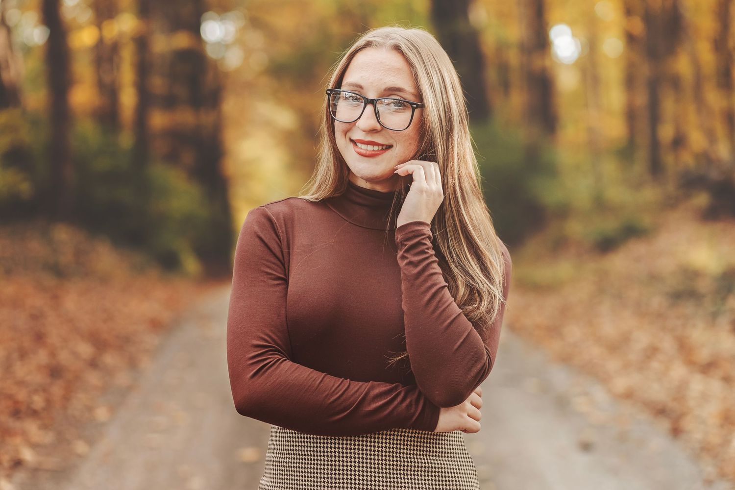Young blonde woman in glasses, brown turtleneck, and mini skirt posing on a forest path in autumn.
