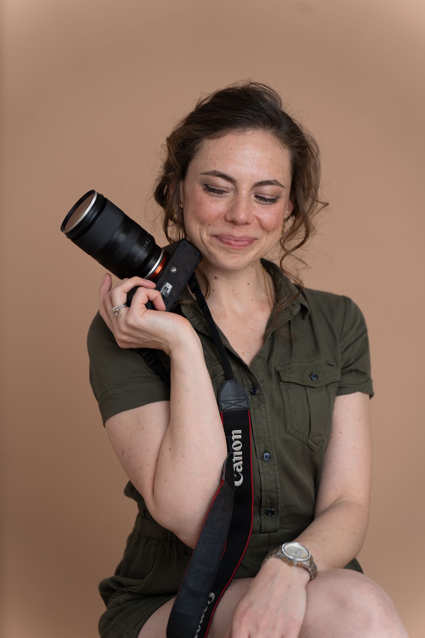 Young woman in olive dress sitting on wooden stool holding Canon DSLR camera, smiling softly against beige background.