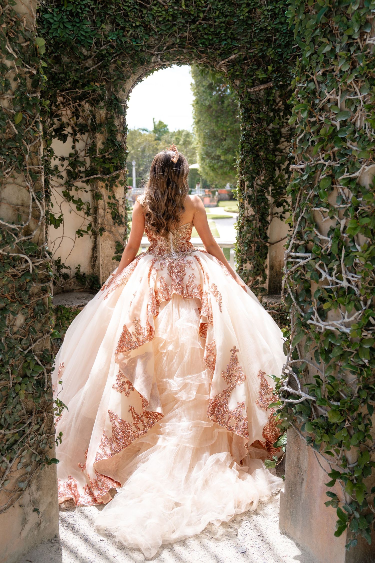 Young woman in elegant blush ballgown with rose gold embroidery stands beneath ivy-covered stone archway.