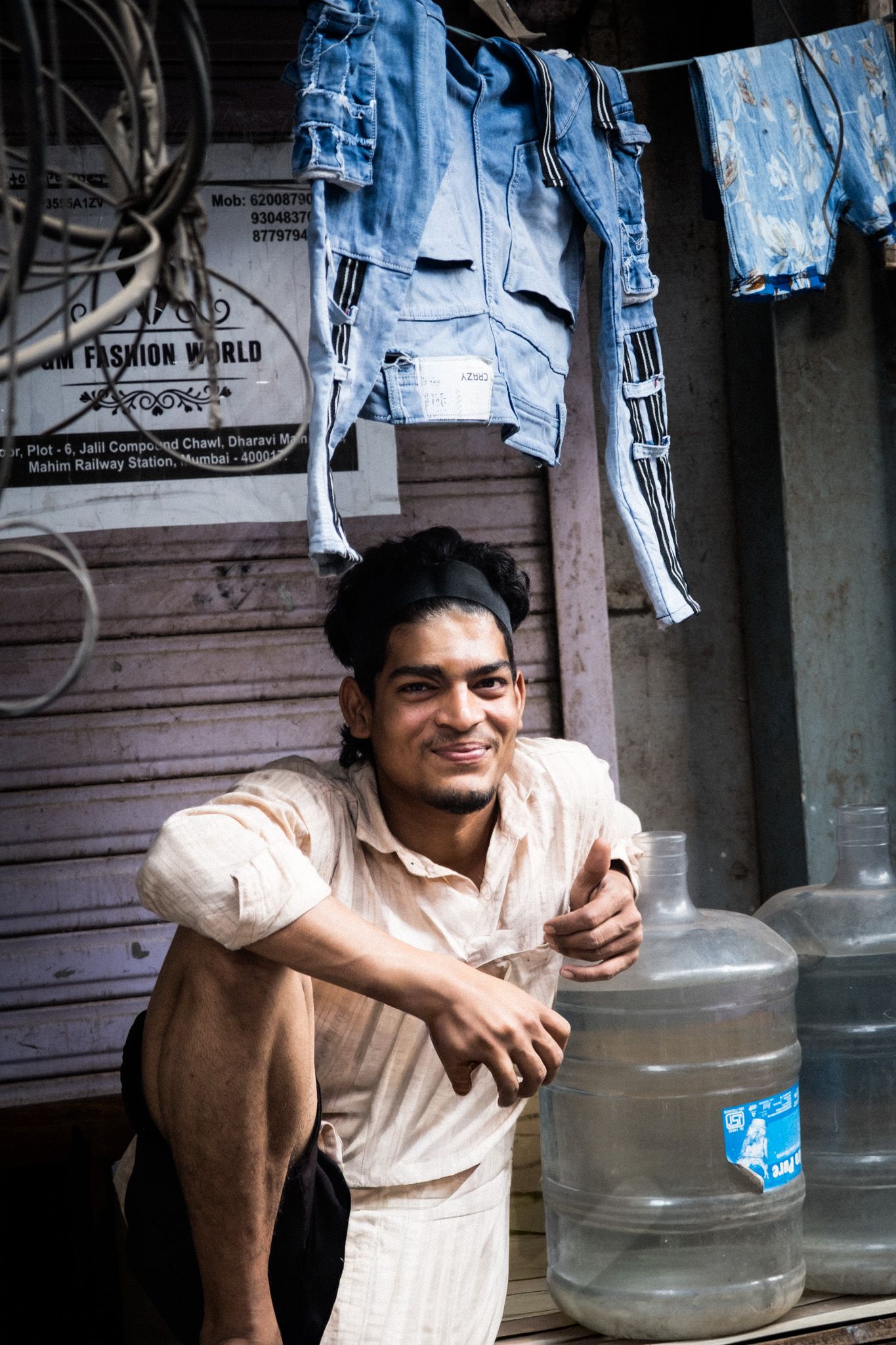A smiling young Indian man sits relaxed outside a small shop with jeans hanging above and water jugs nearby.