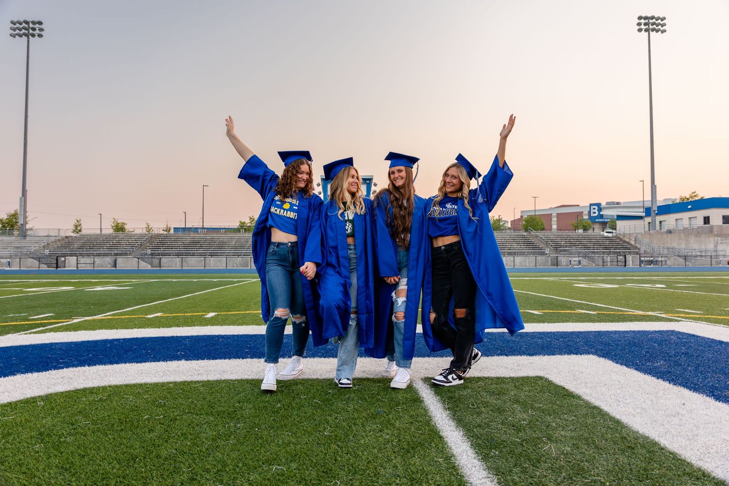 Group of graduates in blue caps and gowns celebrate on a football field during sunset.