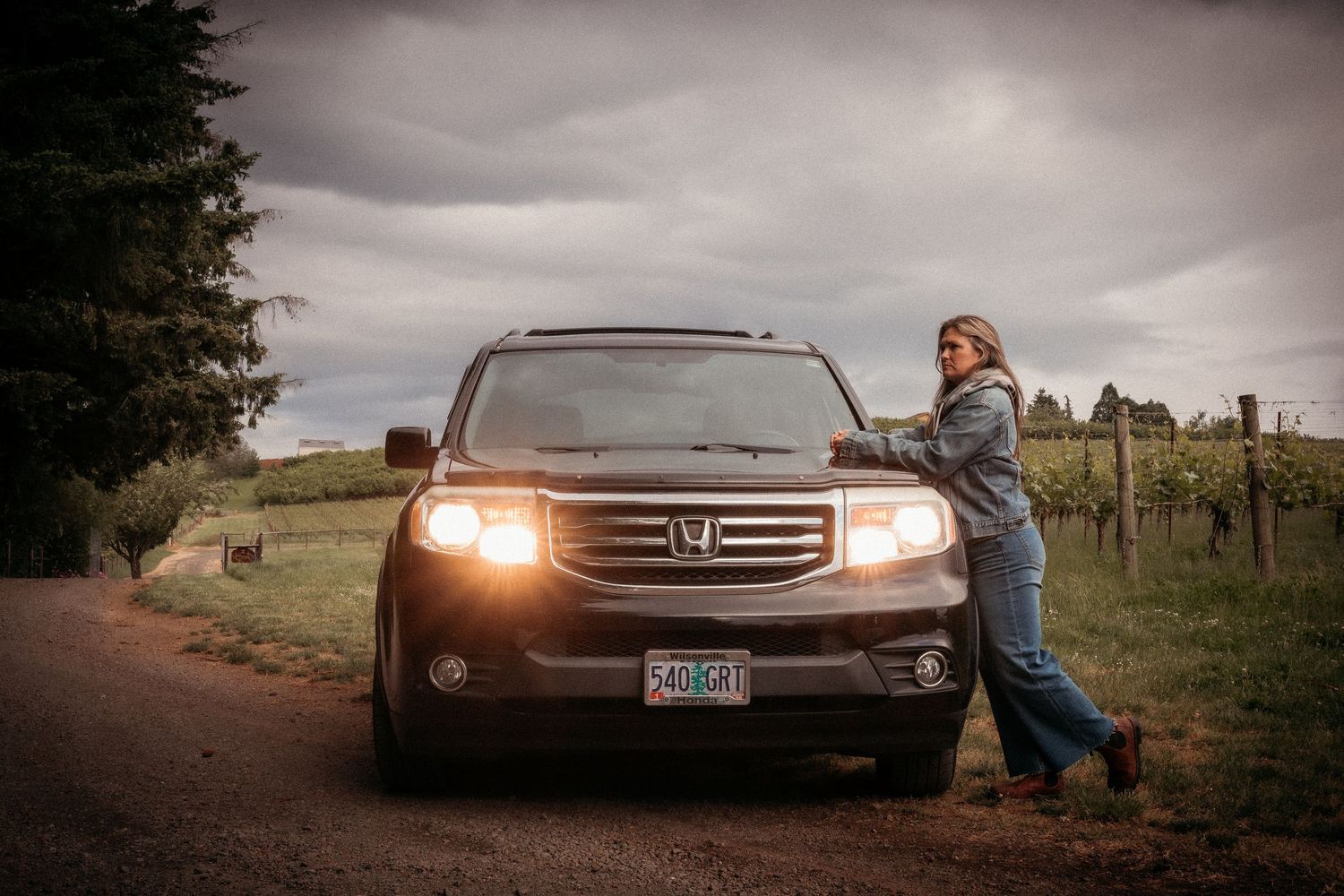 Woman leaning on a Honda SUV with headlights on, parked near a vineyard under a dramatic cloudy sky.