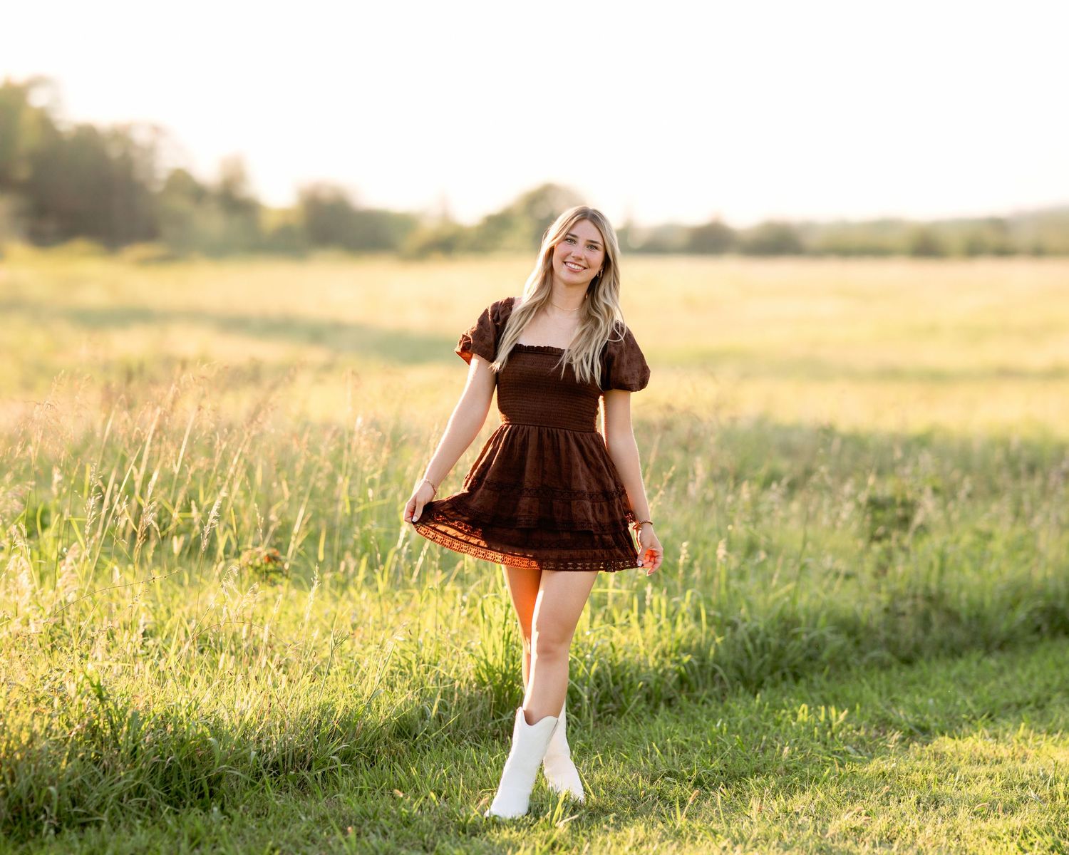 A smiling young woman in a light blue dress leans on a wooden fence in a lush green park setting.