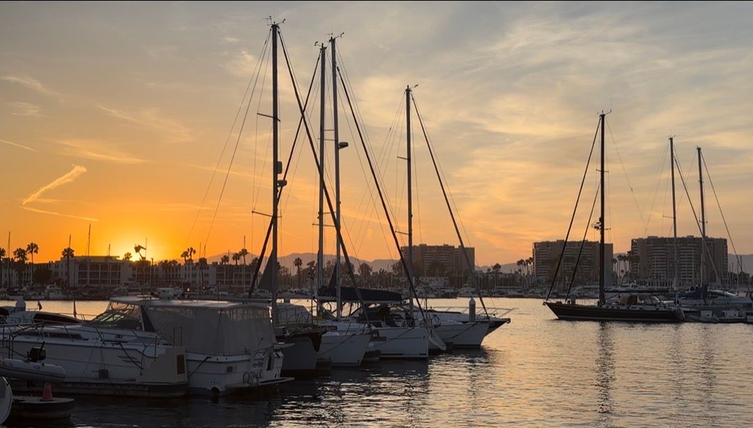Sailboats docked at a marina during a stunning golden sunset with warm orange skies reflecting on calm water.