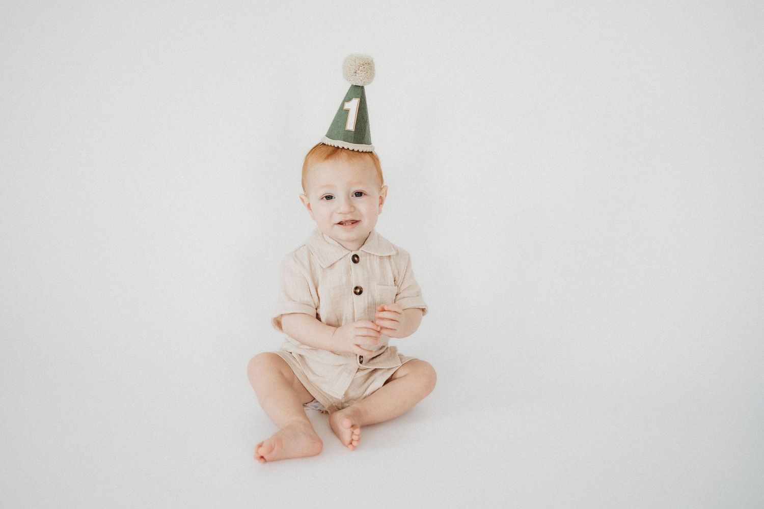 A red-haired baby in a beige romper sits wearing a green number one birthday party hat on a white background.